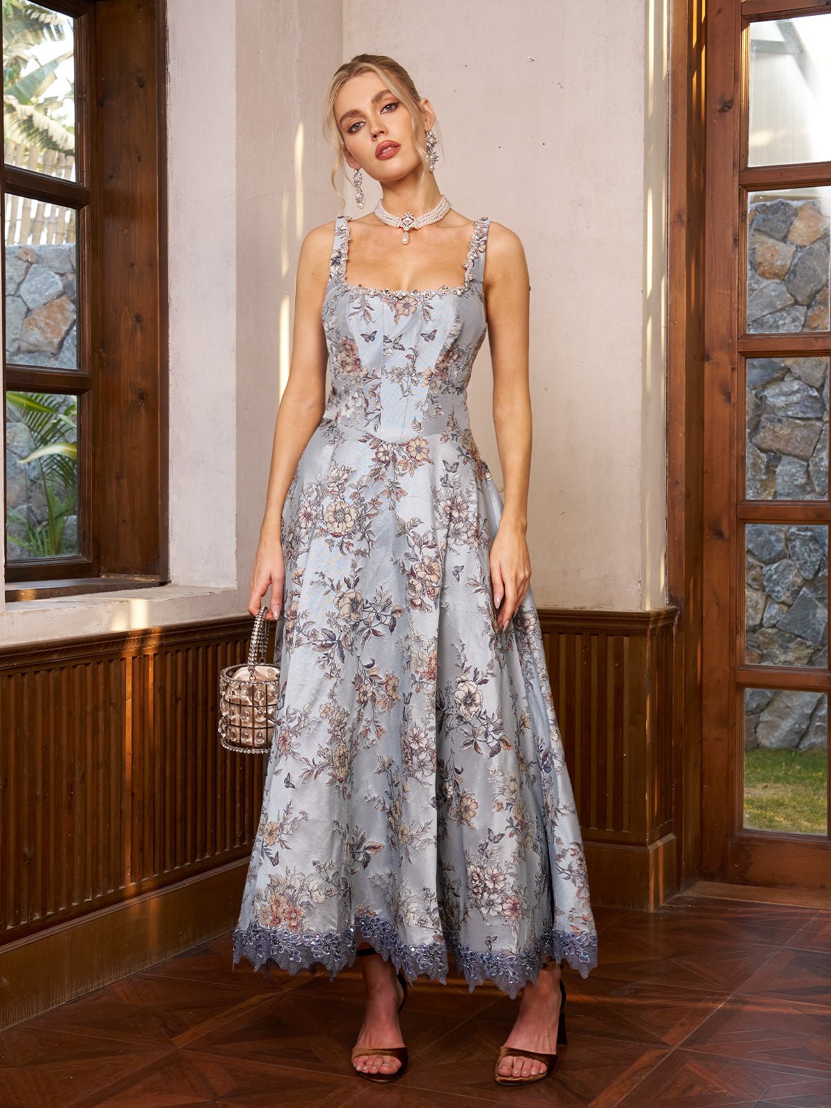 A woman stands indoors by windows in the Social Queen Jacquard Strapless Dress with Beaded Detail, pairing it with a small handbag, earrings, and heels—an elegant wedding guest look.