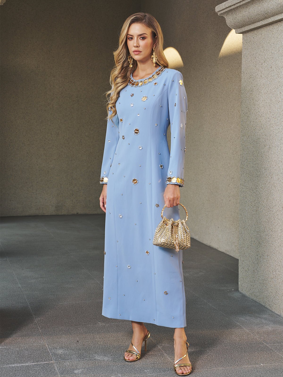 A woman indoors wears the Social Queen Jewel-Beaded Gold-Trim Dress, a long, light blue gown with intricate jewel embellishments—styled with a gold handbag and gold sandals, it's the perfect choice for a stunning wedding guest look.