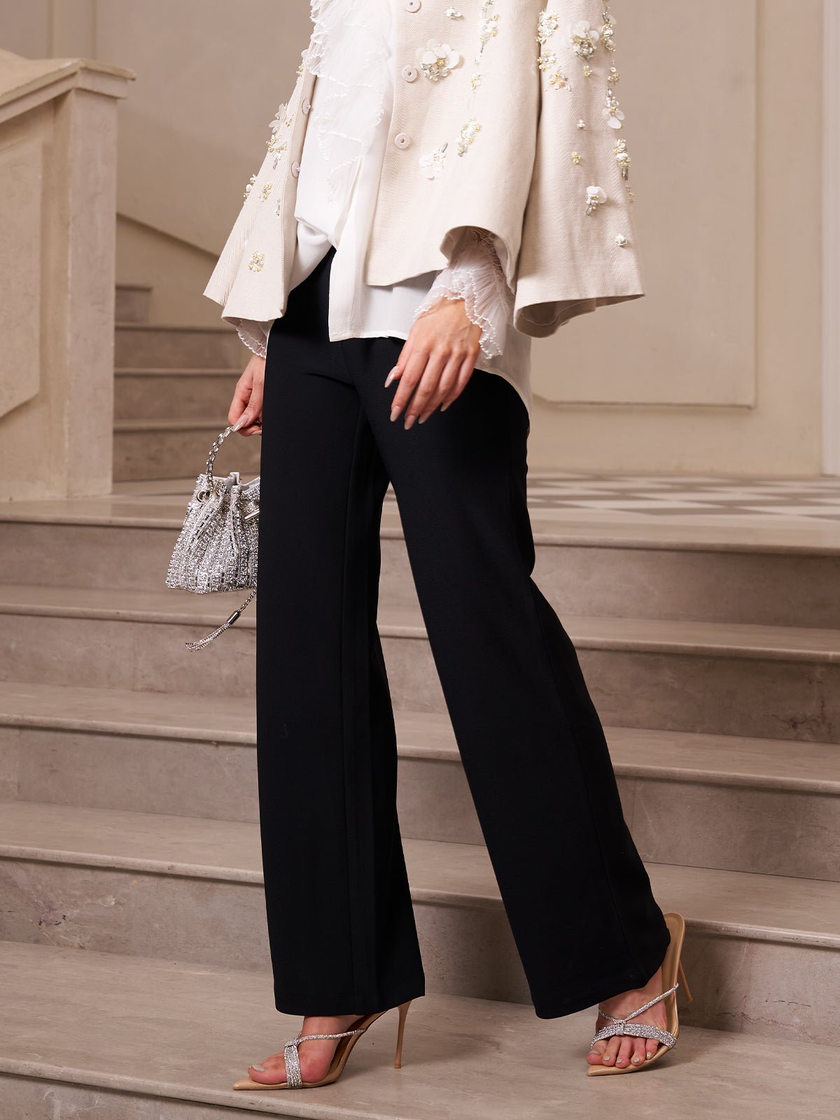 Person wearing a cream embellished jacket, white blouse, Social Queen Lace-Trim Straight-Leg Trousers, and heeled sandals, holding a small handbag while standing on indoor stairs.