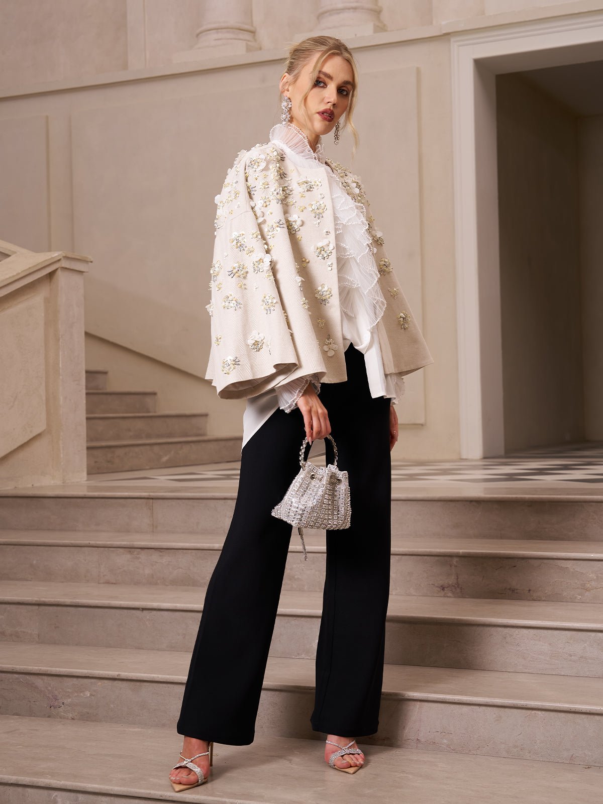 A woman stands on stone stairs wearing a white embellished jacket, white ruffled blouse, Social Queen Lace-Trim Straight-Leg Trousers, silver heels, and holding a small pearl handbag.