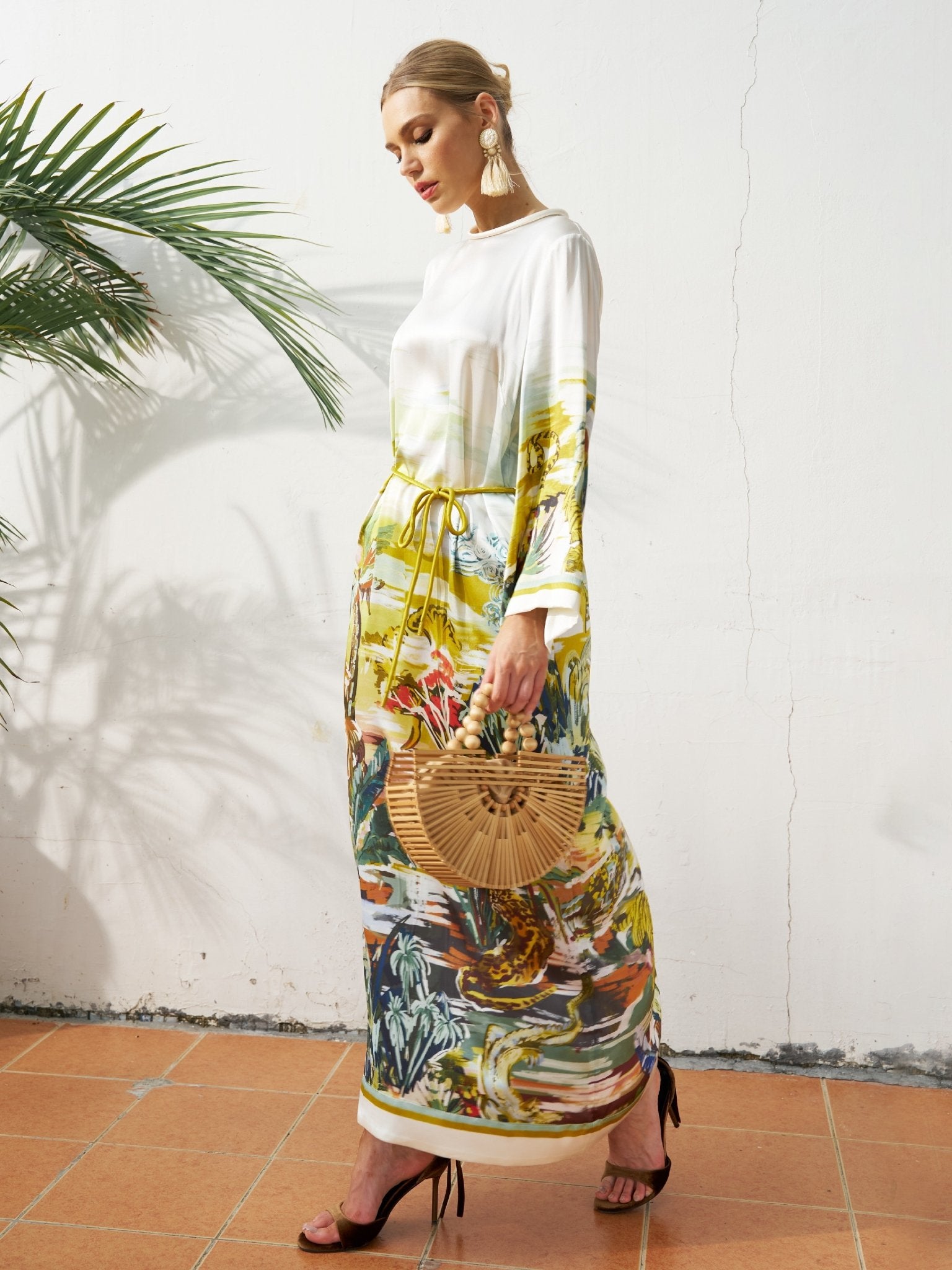 A woman stands indoors wearing the Social Queen Landscape Print Satin Silk Dress, paired with a wooden handbag, statement earrings, and black heels, with a potted palm in the background.