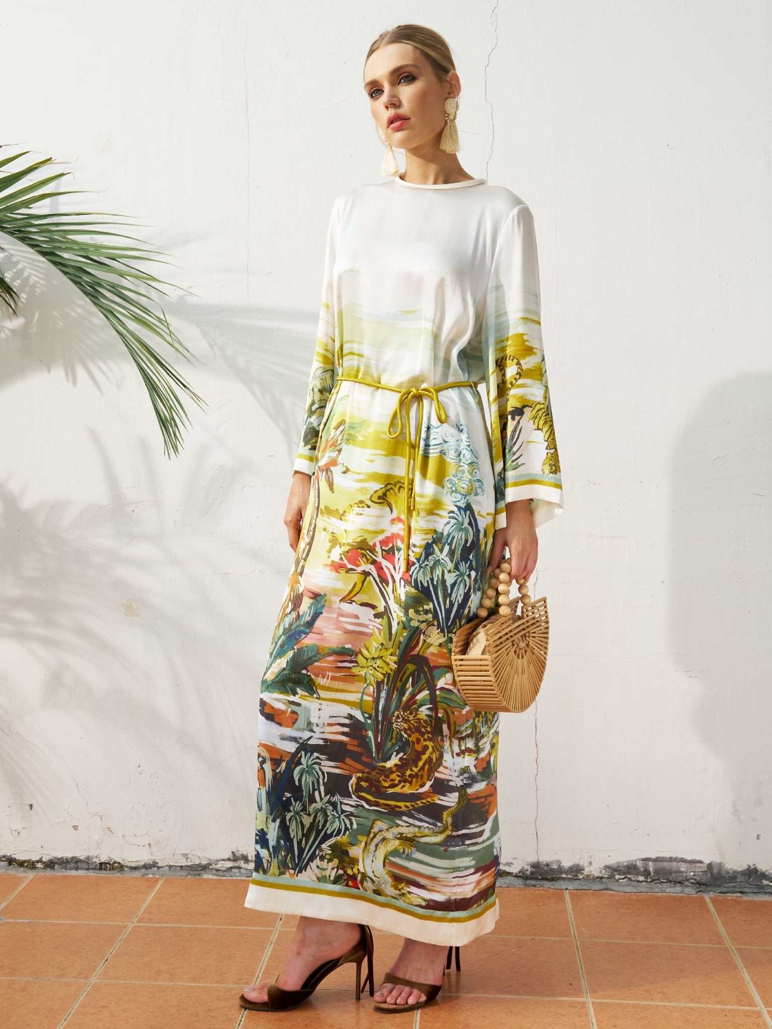 A woman stands against a white wall wearing the Social Queen Landscape Print Satin Silk Dress with a relaxed fit and waist tie, holding a woven handbag; a palm leaf appears on the left.