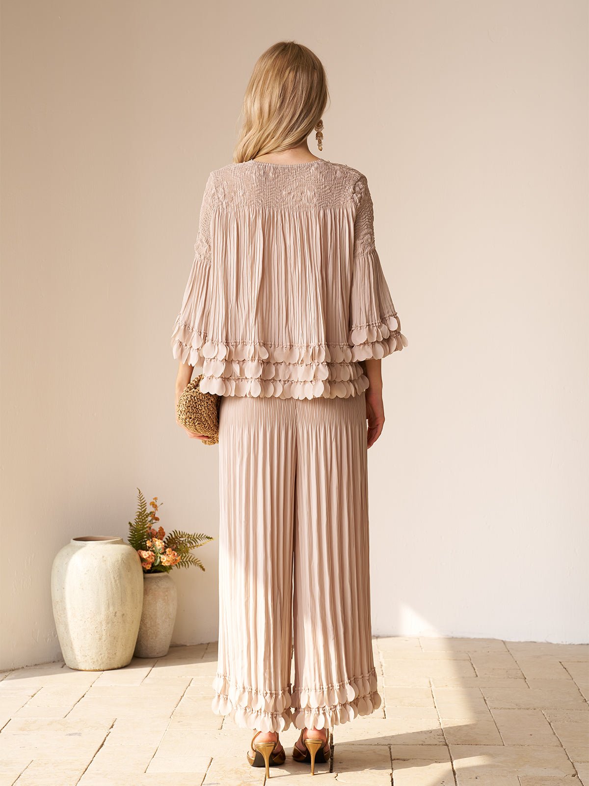 A woman with blonde hair faces away, wearing Social Queen's Layered Petal Beaded Pleated Set—wide-leg pants and textured details—holding a clutch. A nearby vase of flowers completes this chic choice for a wedding guest outfit.