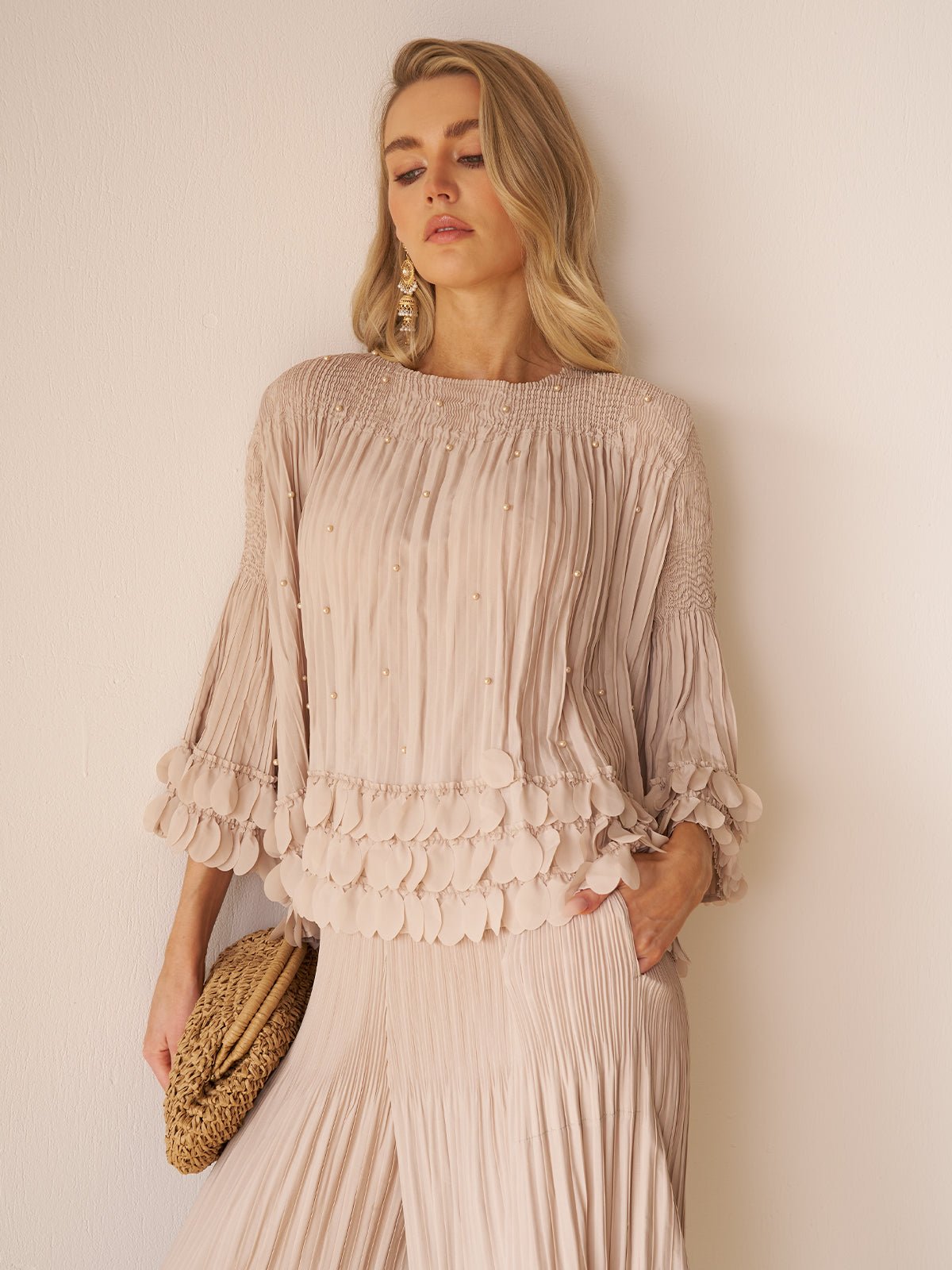 A woman models the Social Queen Layered Petal Beaded Pleated Set—a chic beige ensemble with wide sleeves and decorative trim—styled with a woven bag, perfect for wedding guests, against a plain light background.