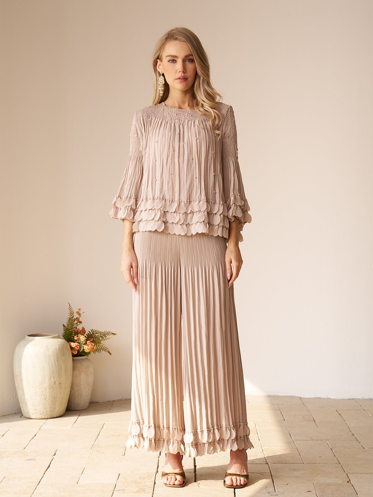 A woman stands indoors in the Social Queen Layered Petal Beaded Pleated Set—light beige with scalloped edges and wide-leg pants—paired with neutral sandals and statement earrings. A ceramic vase with flowers is in the background.