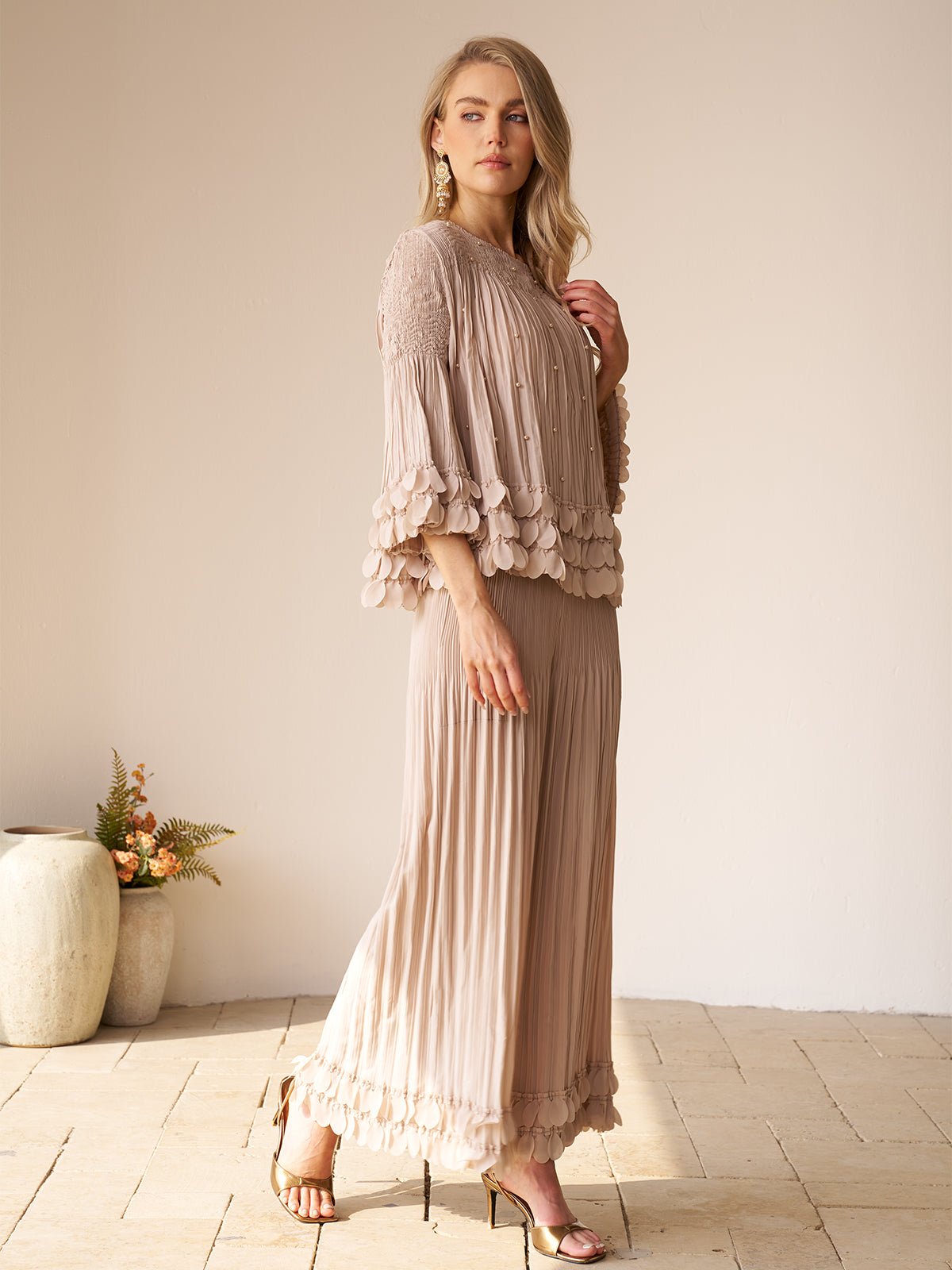 A woman stands indoors on a tiled floor by a ceramic vase with flowers, wearing the Social Queen Layered Petal Beaded Pleated Set featuring textured sleeves and hem in light beige.