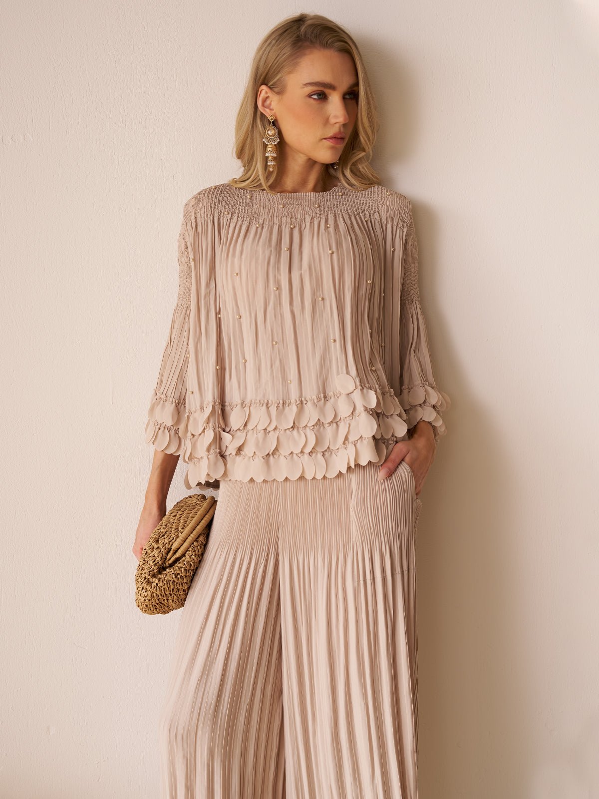 A woman stands against a white wall in Social Queen's Layered Petal Beaded Pleated Set—a light beige, wide-leg two-piece—styled with a woven clutch and statement earrings for an effortlessly chic wedding guest look.