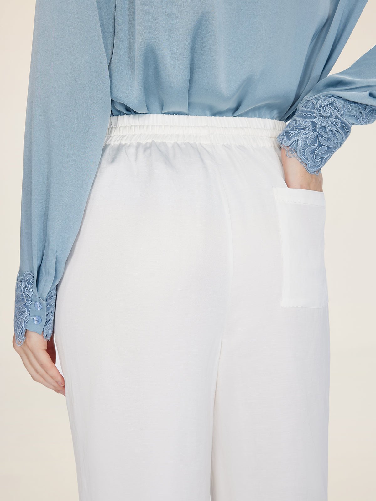 A person wears Social Queen Linen Straight-Leg Pants—white, breathable linen with an elastic waistband and back pocket—paired with a light blue long-sleeve blouse featuring lace cuffs.