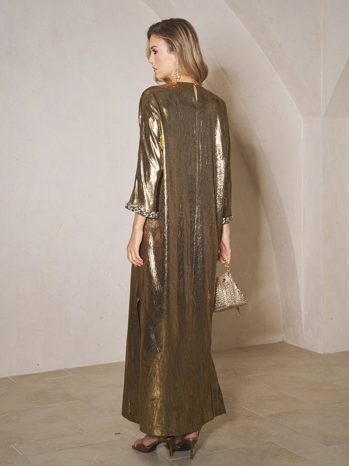 A woman in the Social Queen Metallic Crystal-Embellished Slit Dress and heels stands indoors, facing away, holding a small handbag.