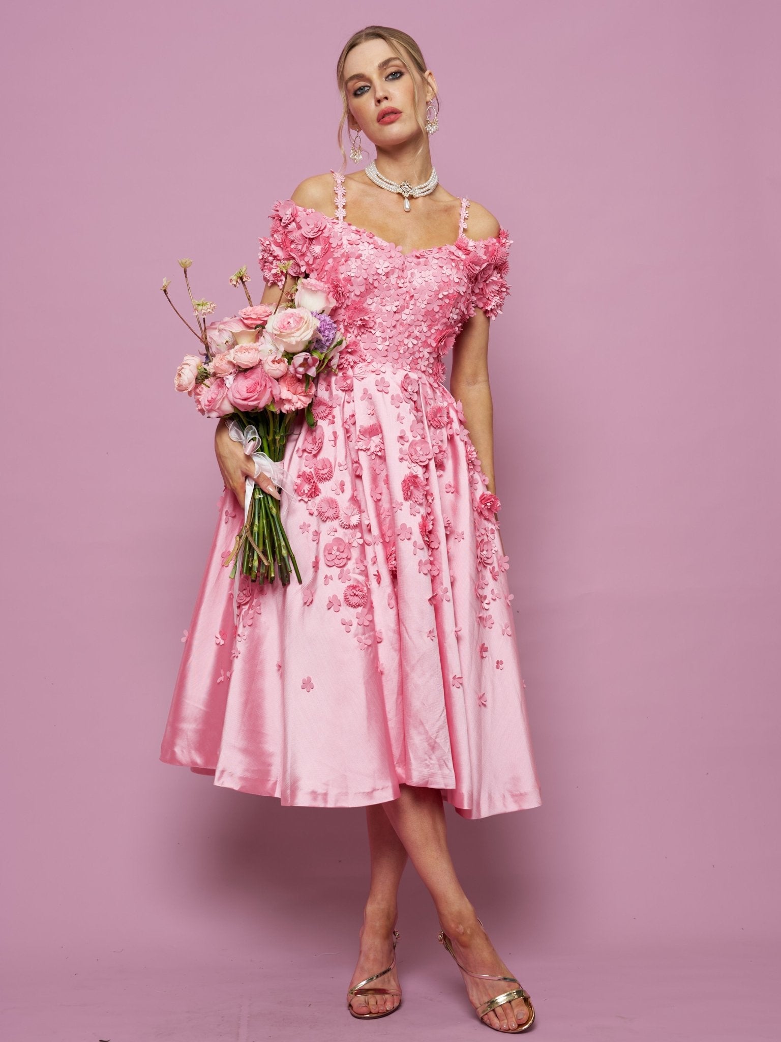 A woman in the Social Queen Off-Shoulder Floral Satin Gown holds a bouquet of pink flowers, standing against a pink background.