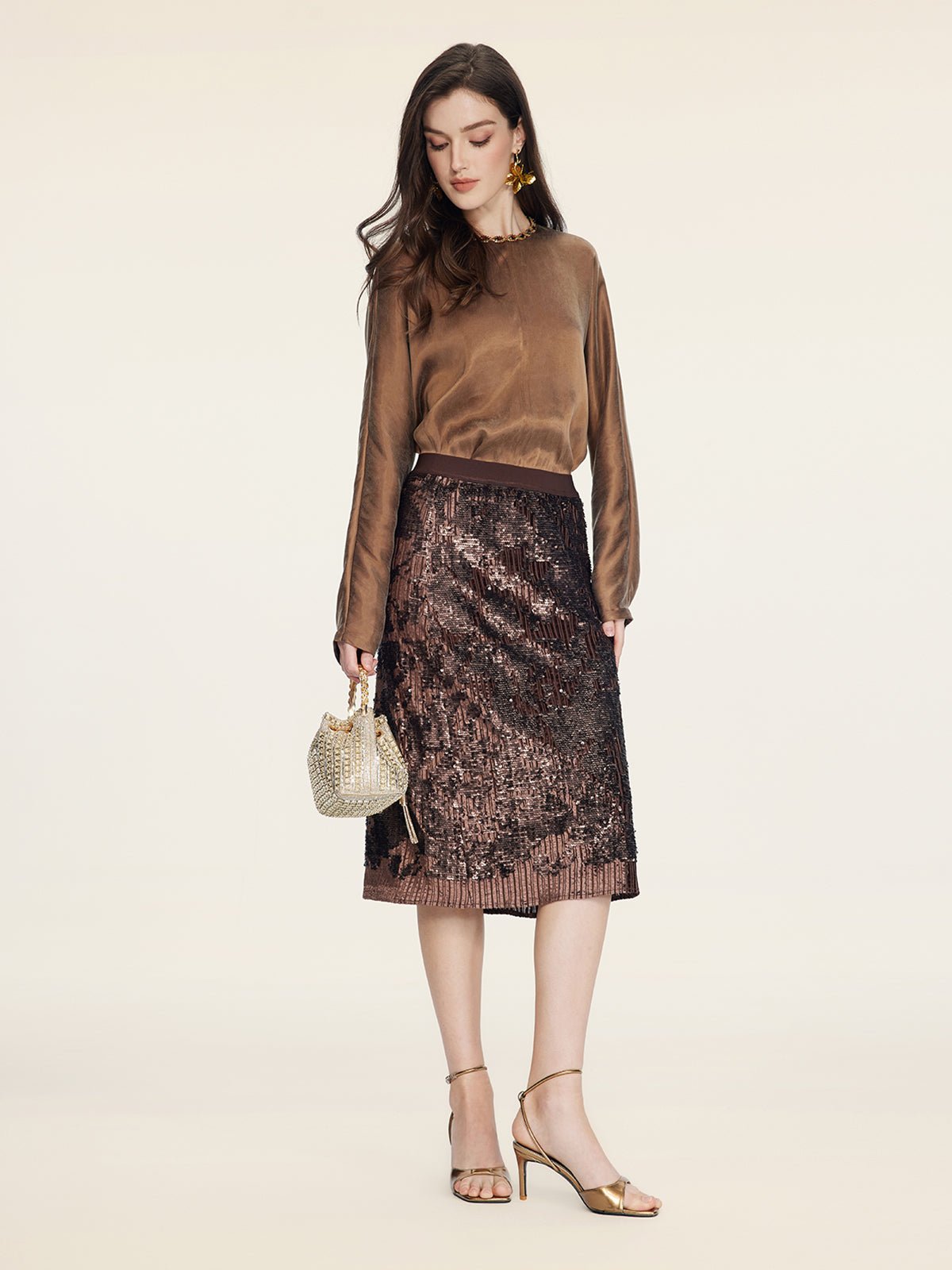 A woman stands against a plain light background wearing a brown long-sleeve blouse, gold heels, a small gold purse, and the Social Queen Ombre Sequin Velvet Skirt.