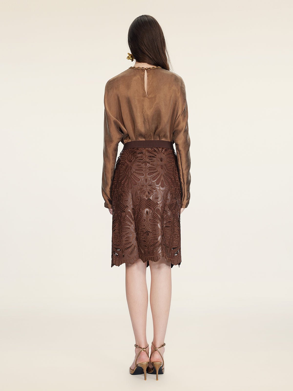 A woman faces away in a sheer brown blouse and the Social Queen Openwork Embroidered Leather Skirt with hand-embroidered details and a wavy hem, paired with heeled sandals, set against an off-white background.