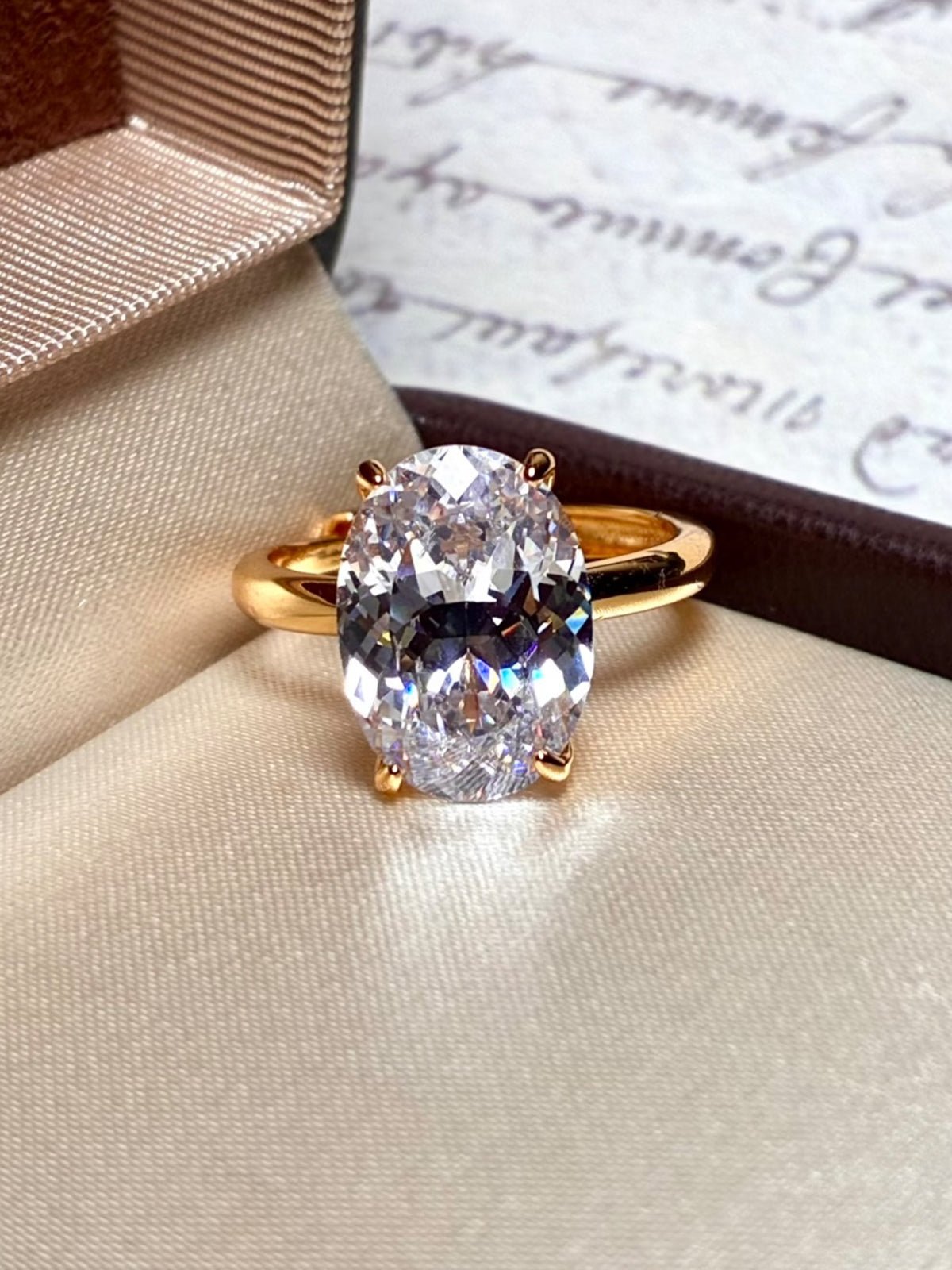 The Social Queen Oval Crystal Ring, featuring a gold-tone adjustable band and an oval-cut clear crystal, is showcased in an open ring box with handwritten paper in the background.