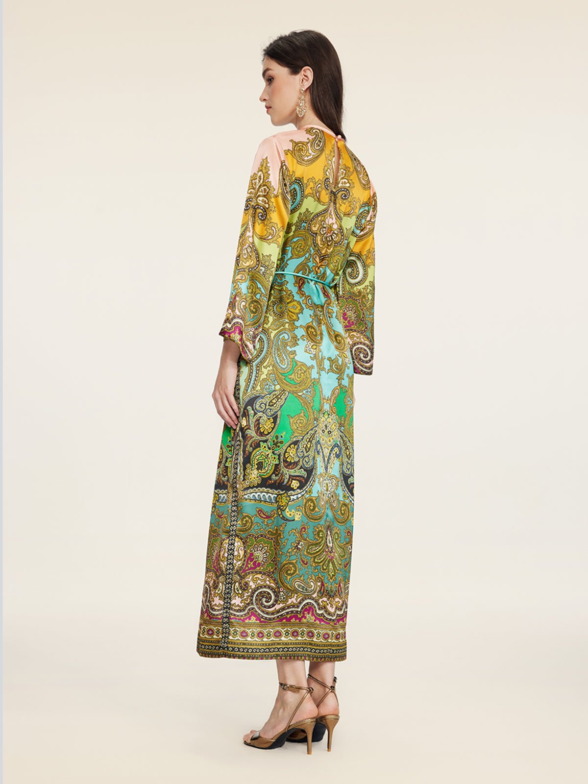 A woman stands sideways and faces away, wearing the Social Queen Paisley Printed Silk Satin Dress with long sleeves and heeled sandals, set against a plain background.