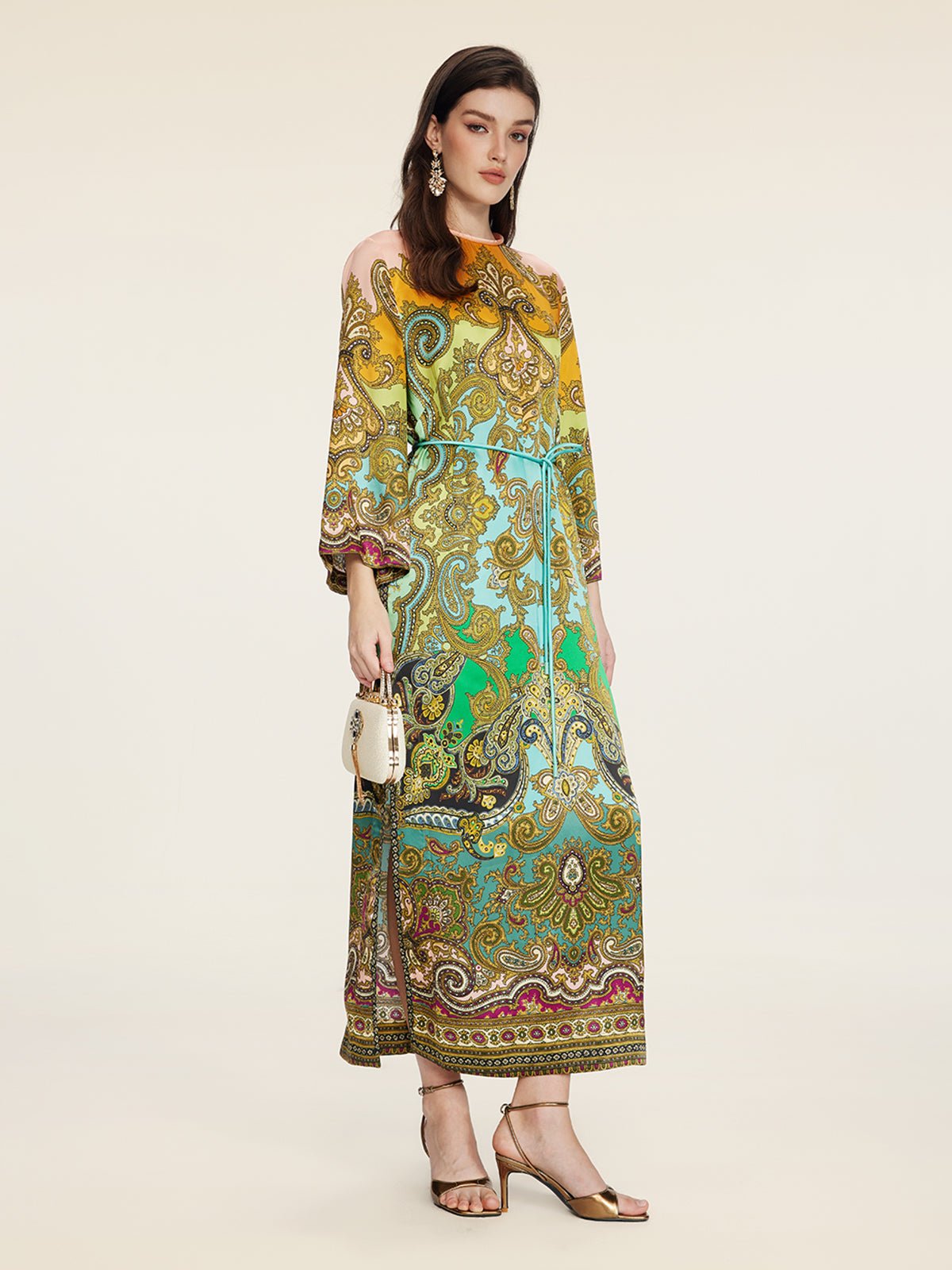 A woman wears the Social Queen Paisley Printed Silk Satin Dress in yellow, green, and brown, paired with a small white handbag and gold sandals.