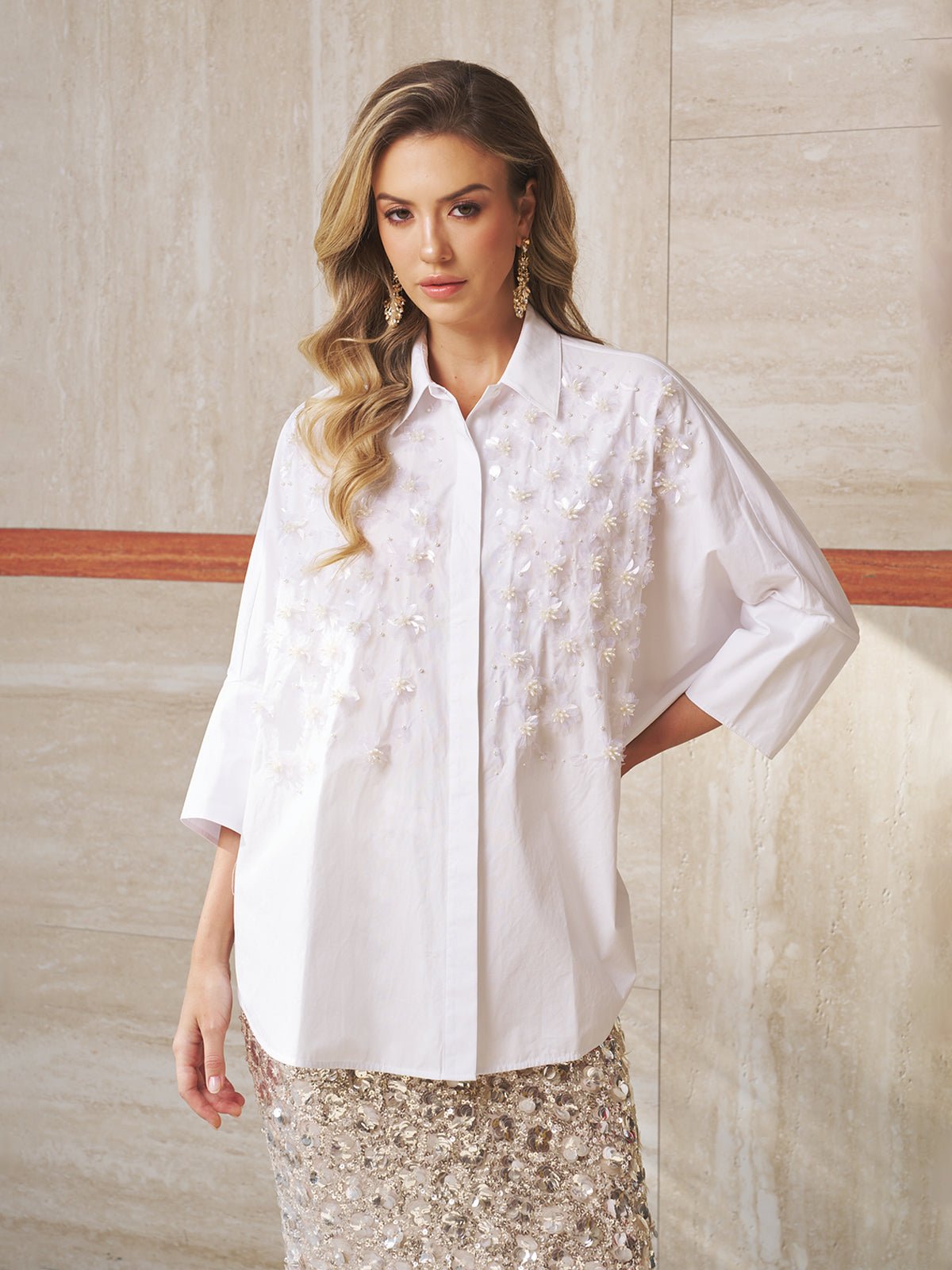 A woman wears the Pearl-Embellished Floral Blouse by Social Queen with a sequined skirt, creating an elegant look perfect for a wedding guest, as she stands against a marble wall.