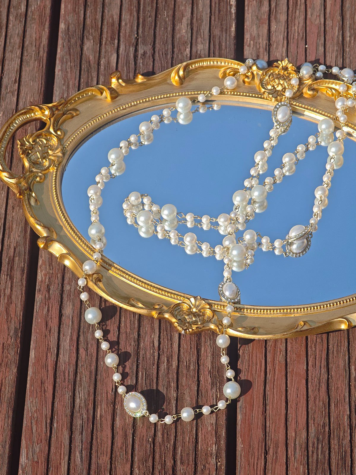 A Social Queen Pearl Necklace with Crystal Accents is draped across a gold-framed mirror, set on a wooden surface.