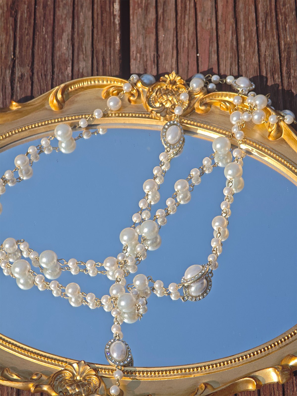 The Social Queen Pearl Necklace with Crystal Accents is displayed on an ornate gold-framed mirror, reflecting a wooden surface in the background.