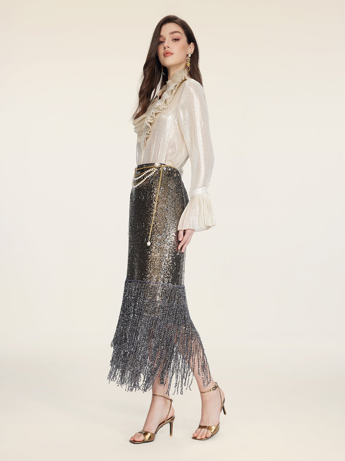 A woman stands sideways against a plain light background, wearing a shimmery long-sleeve blouse, gold heels, gold jewelry, and the Social Queen Pearl-Trim Sequin Fringe Skirt.