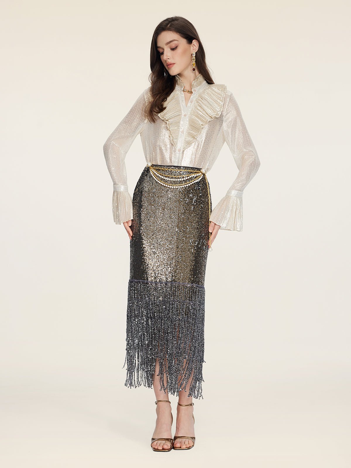 A woman wears the Social Queen Pearl-Trim Sequin Fringe Skirt with a sheer ruffled blouse, gold chain belt, earrings, and high-heeled sandals against a plain background.