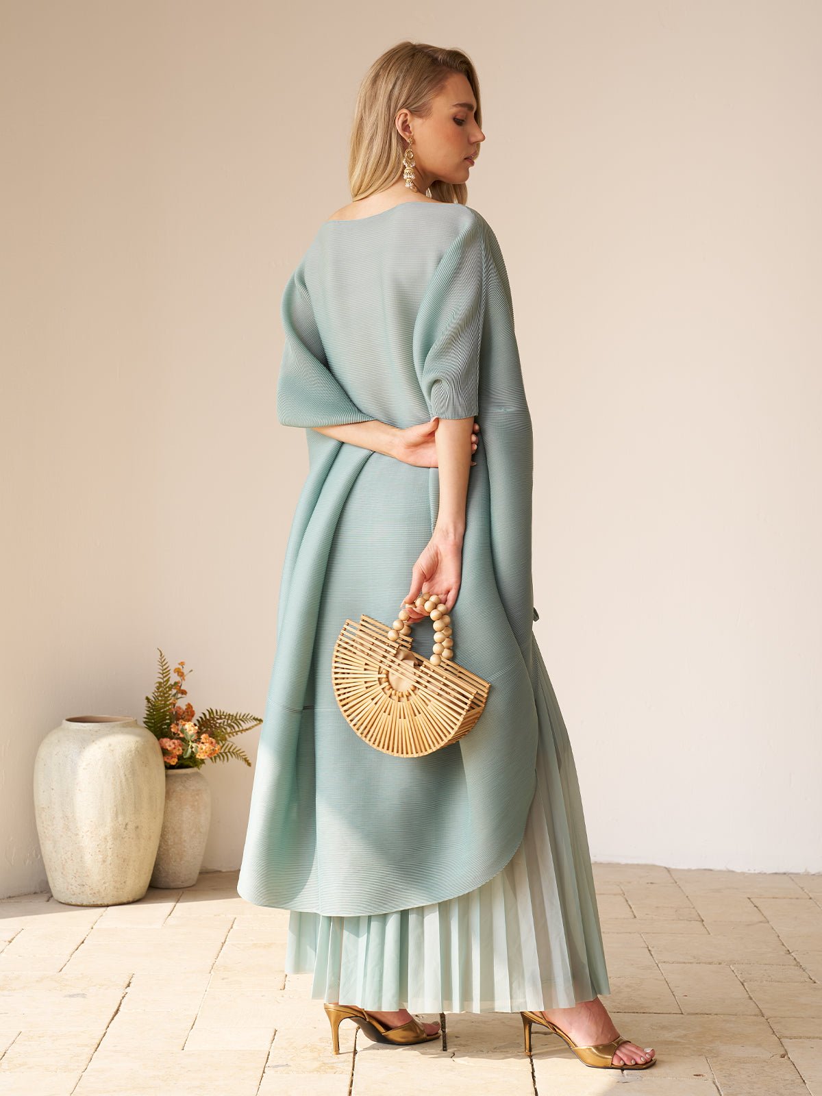 A woman models the Social Queen Pleated Floral Appliqué Set, holding a wooden handbag while standing on tiled flooring with decorative vases and floral arrangements in the background.