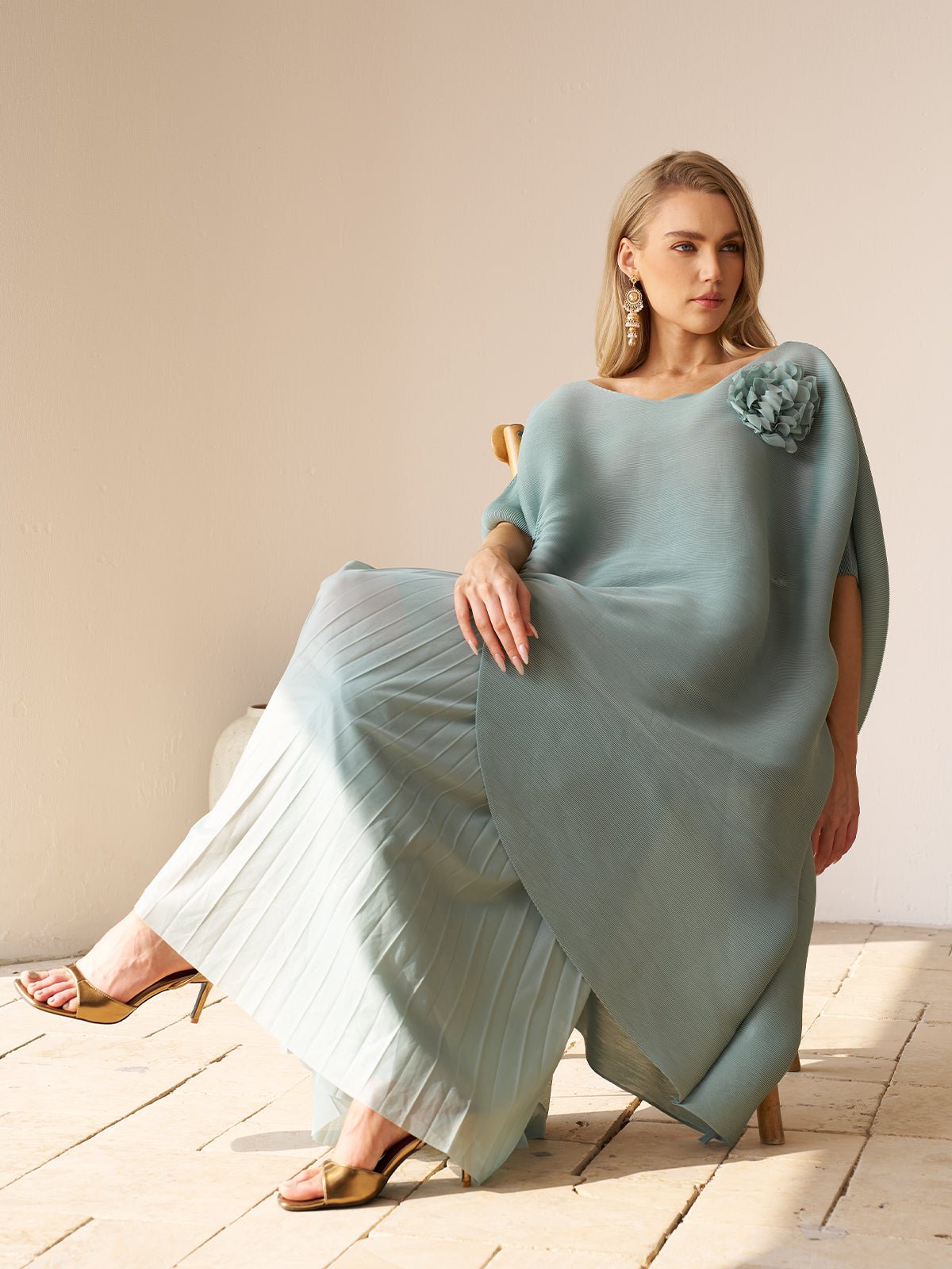 A woman with blonde hair and gold earrings sits on a wooden chair wearing the Social Queen Pleated Floral Appliqué Set—an elegant light blue high-waist chiffon ensemble with 3D floral details—paired with heeled sandals.