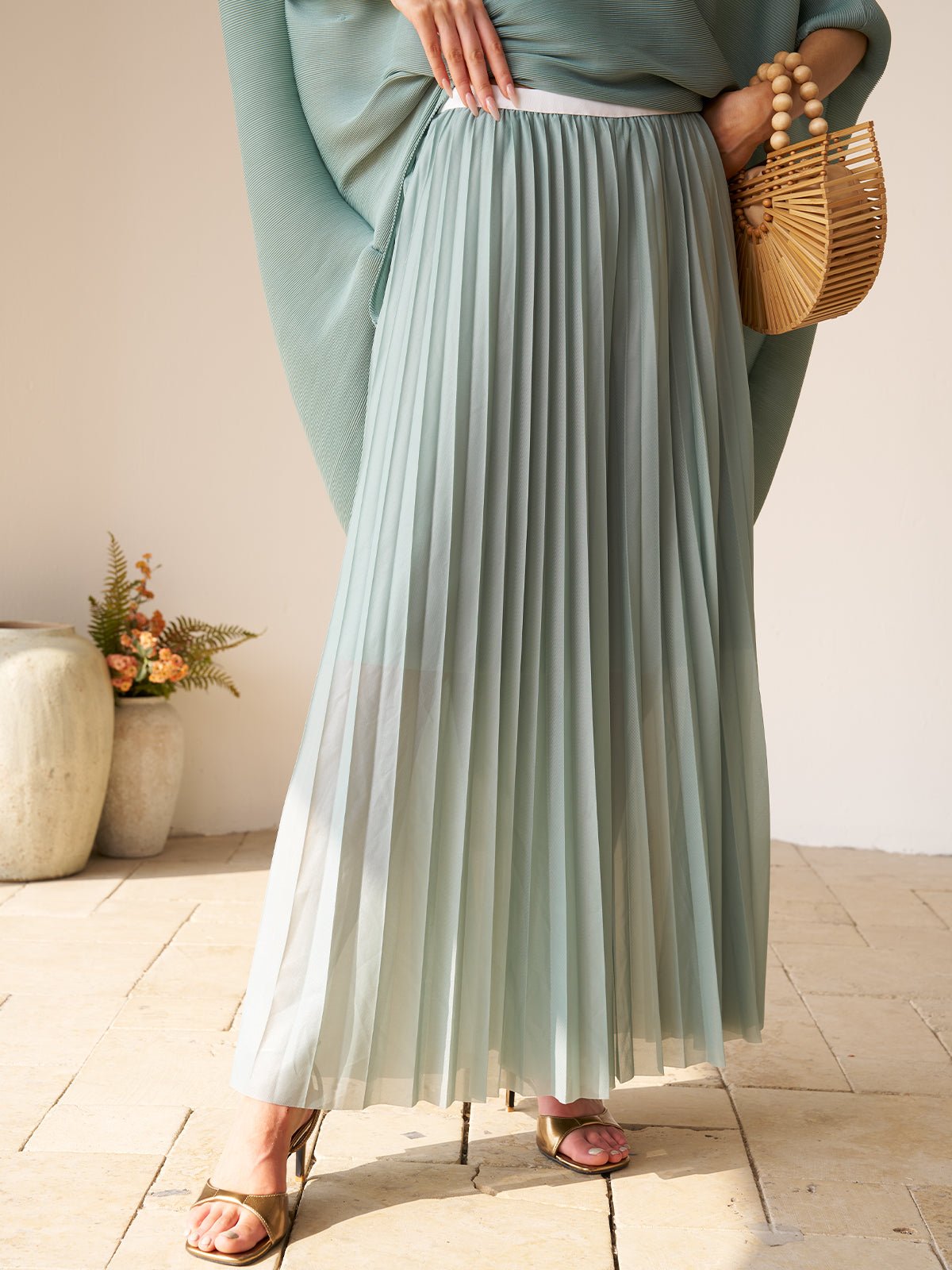Wearing the Social Queen Pleated Floral Appliqué Set and gold sandals, a person holds a woven wooden handbag while standing on tiled flooring beside a ceramic vase with flowers.