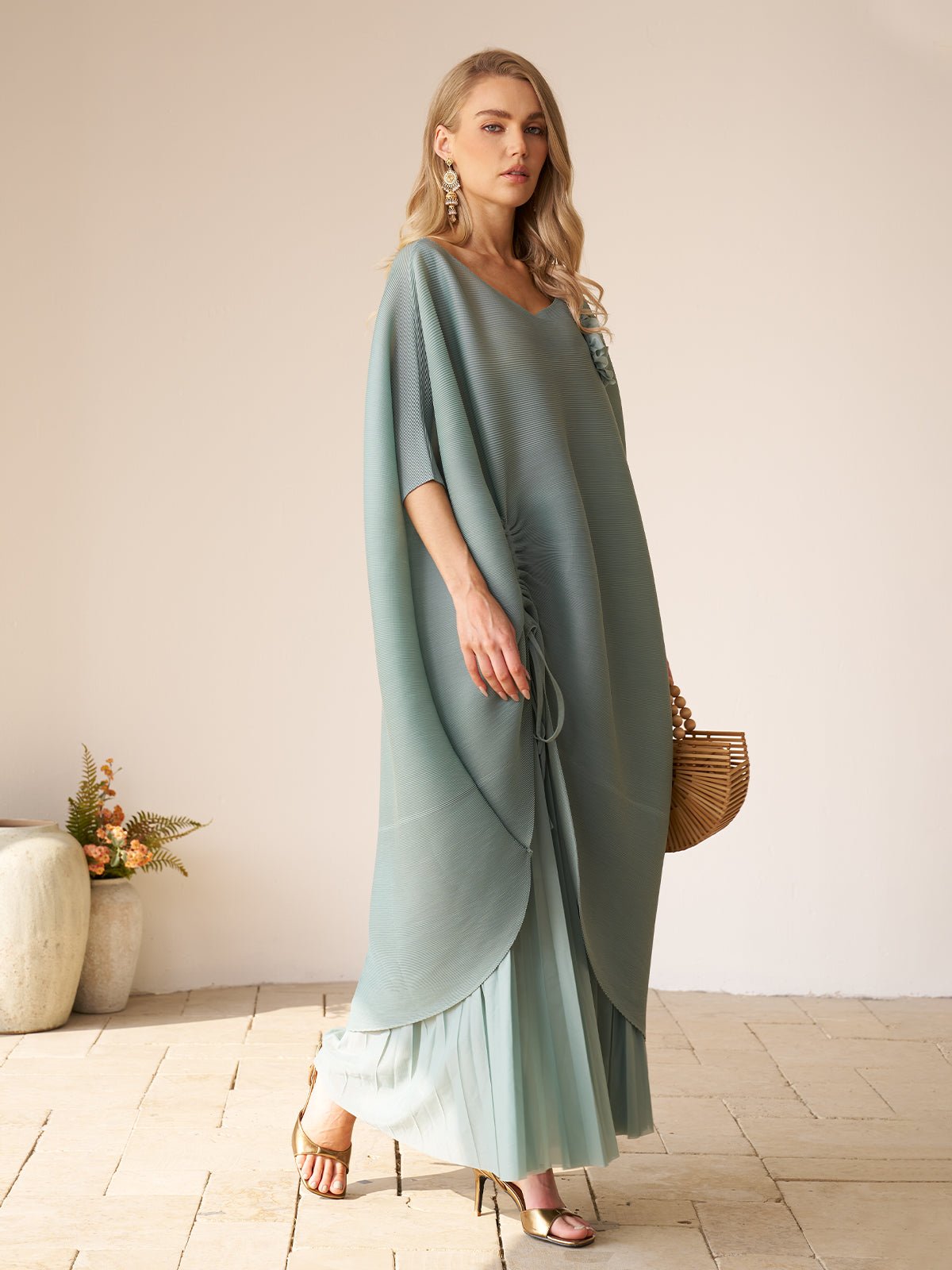 A woman stands indoors wearing the Social Queen Pleated Floral Appliqué Set—loose teal draped top with high-waist pleated pants—holding a wooden bag, with ceramic pots of flowers in the background.