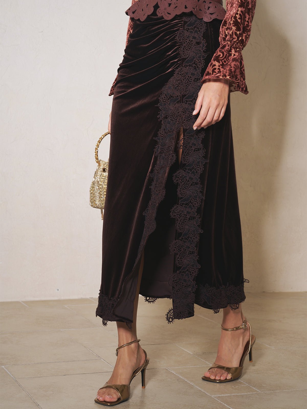 A person wears Social Queen’s Pleated Lace-Panel Velvet Skirt in dark brown with a pleated waist, lace trim, and front slit, styled with a red patterned top, gold sandals, and a small gold bag.