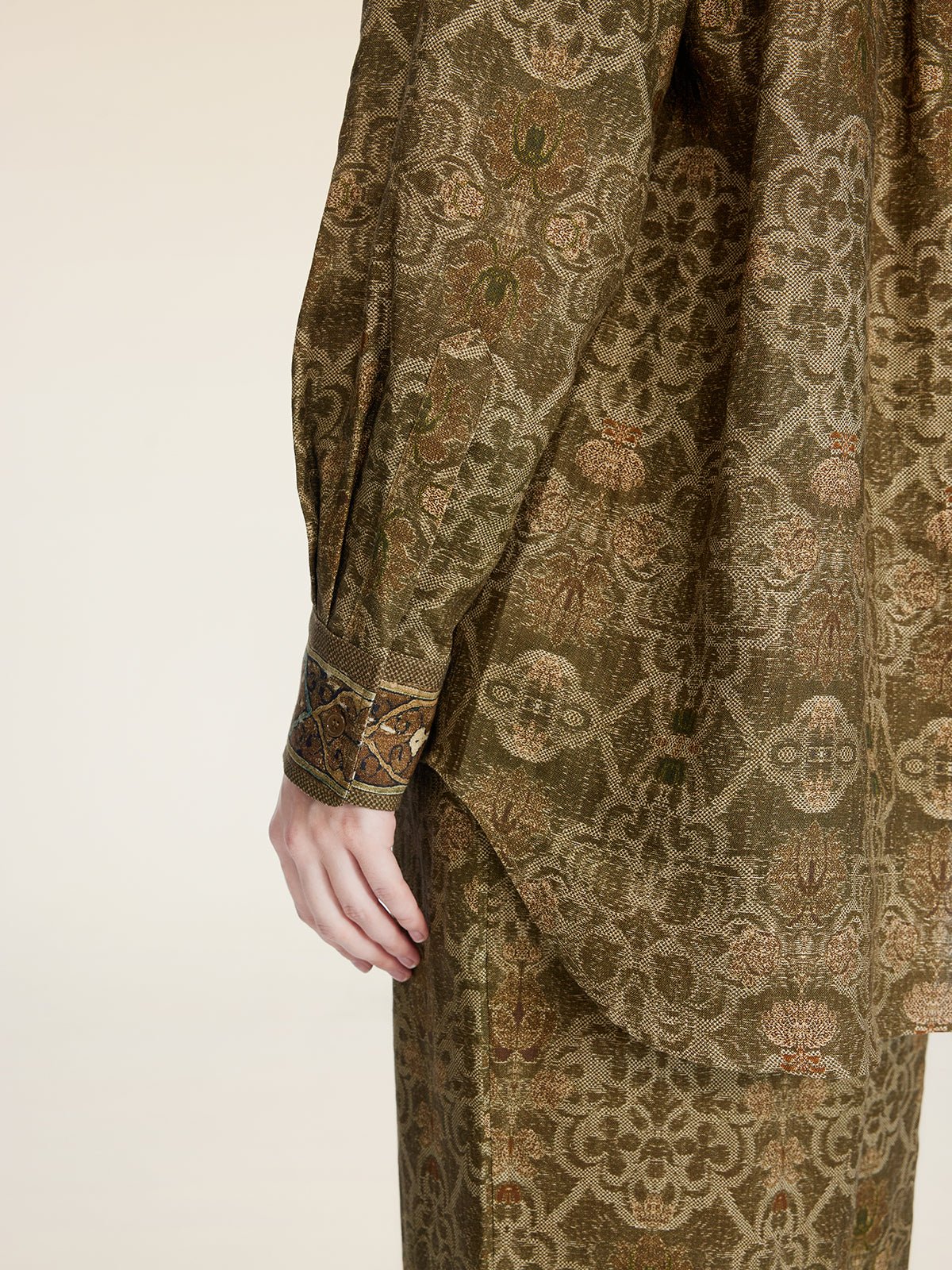 A person models the Social Queen Printed Linen Blouse, featuring long sleeves and an intricate olive green pattern, shown from shoulder to mid-thigh with their hand by their side.
