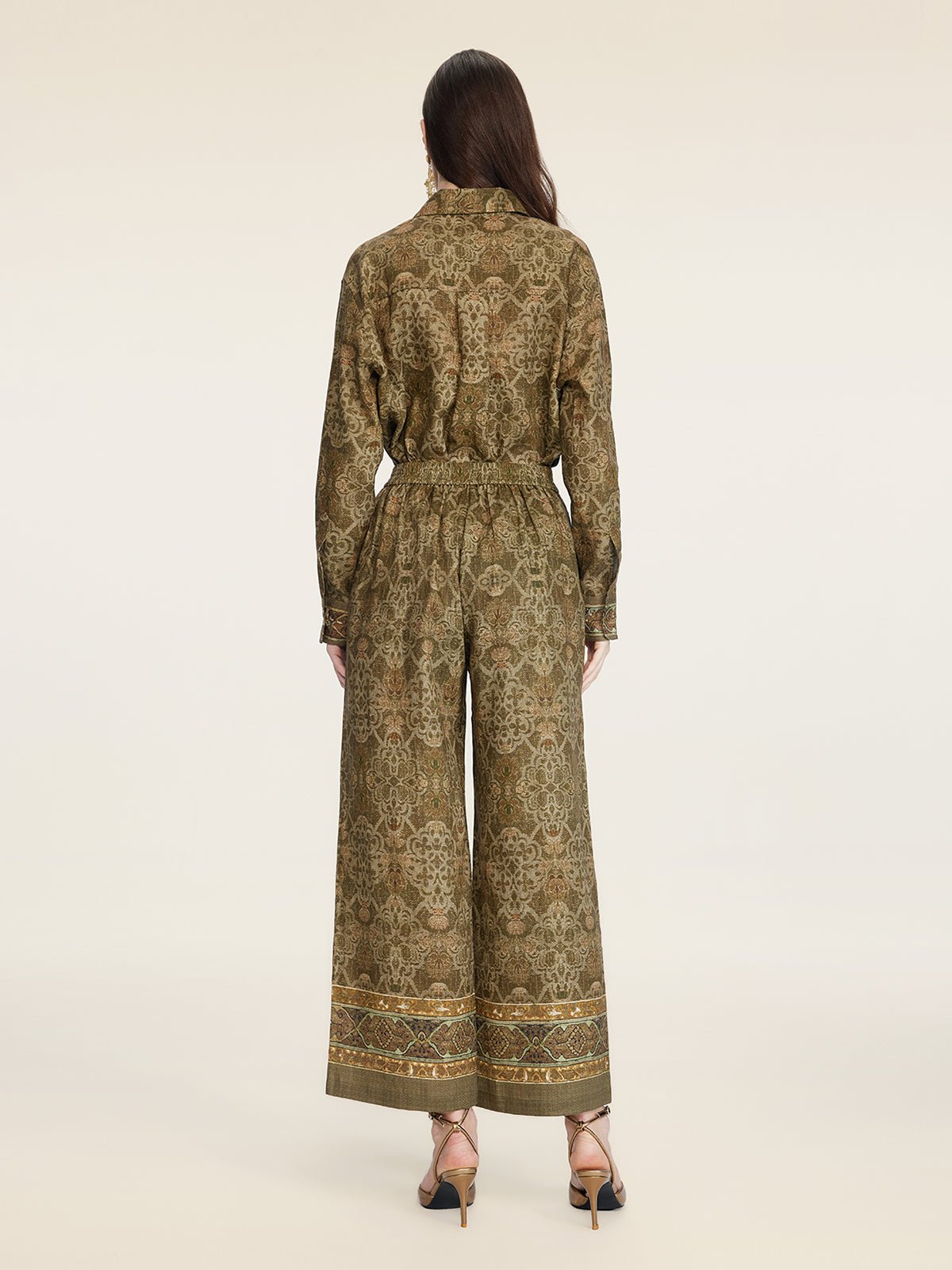 A person stands facing away, wearing Social Queen's Printed Linen Straight-Leg Pants in olive green with gold detailing, paired with a matching patterned long-sleeve top and open-toe heels.