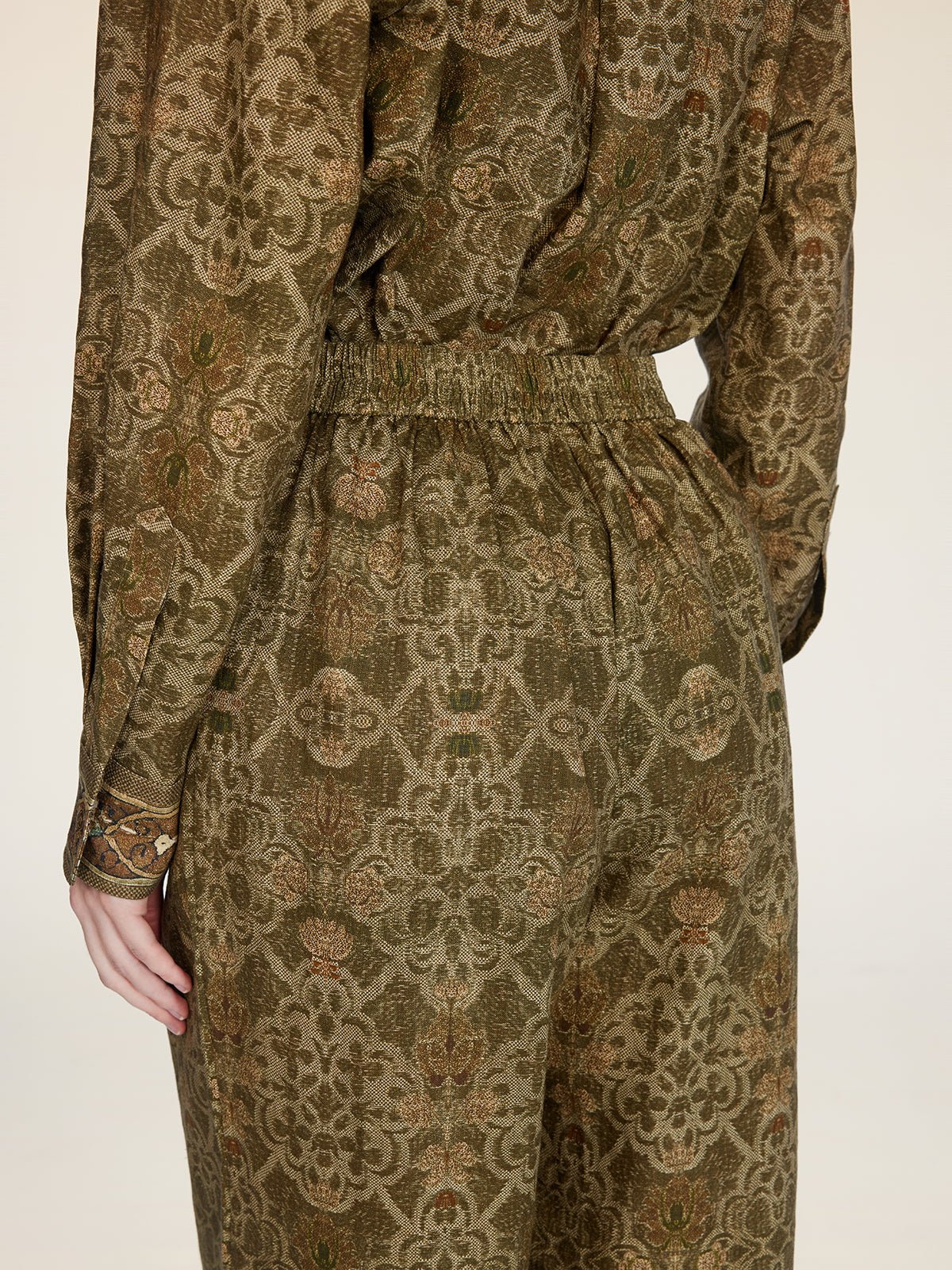 A person is shown from the back wearing Social Queen’s Printed Linen Straight-Leg Pants in olive green and brown with an intricate floral pattern, paired with a coordinating long-sleeve top.