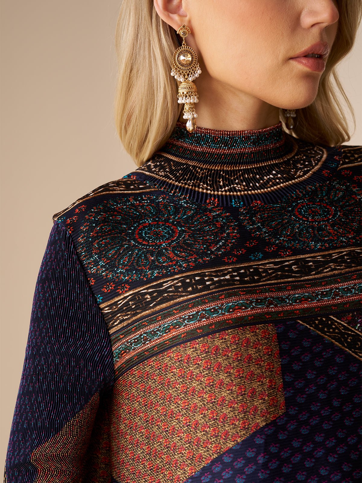A blonde woman wears ornate gold earrings and the Social Queen Retro Ethnic Print Pleated Dress featuring a high neck, multicolored patterns, and intricate designs.
