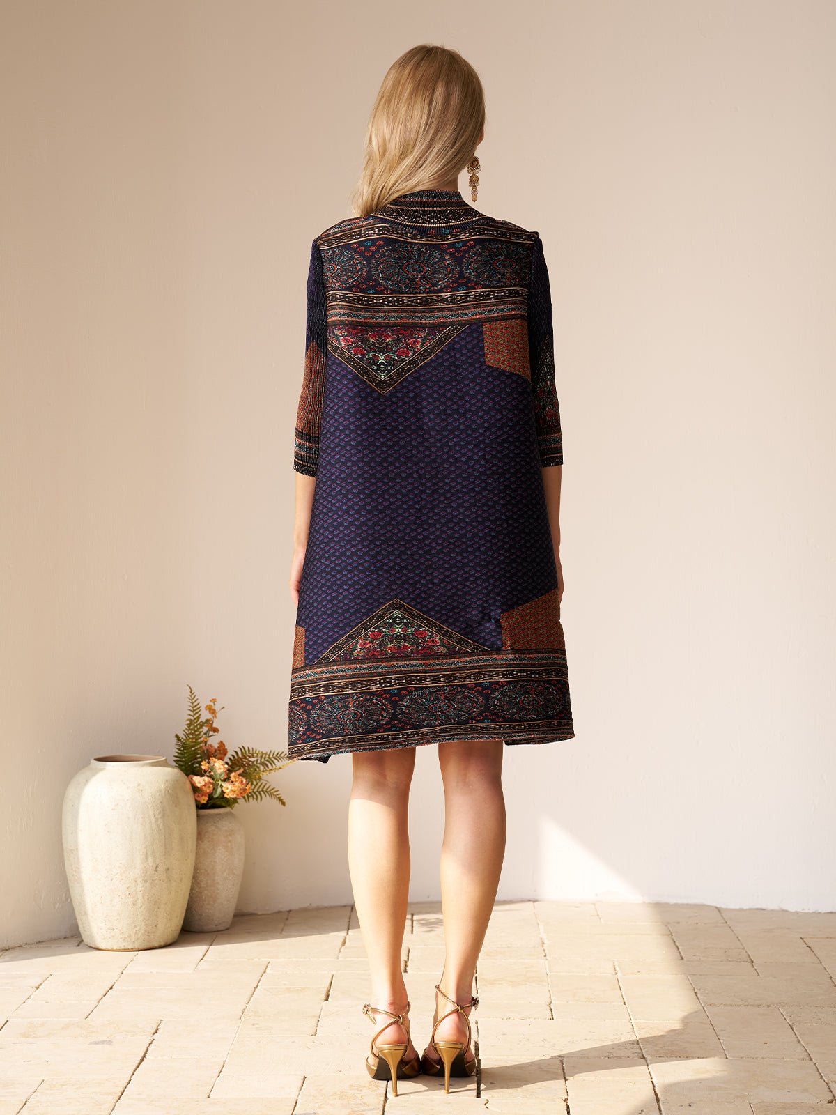 A woman with blonde hair, wearing the Retro Ethnic Print Pleated Dress by Social Queen and heeled sandals, stands indoors facing away from the camera. A vase with flowers is on the floor nearby.