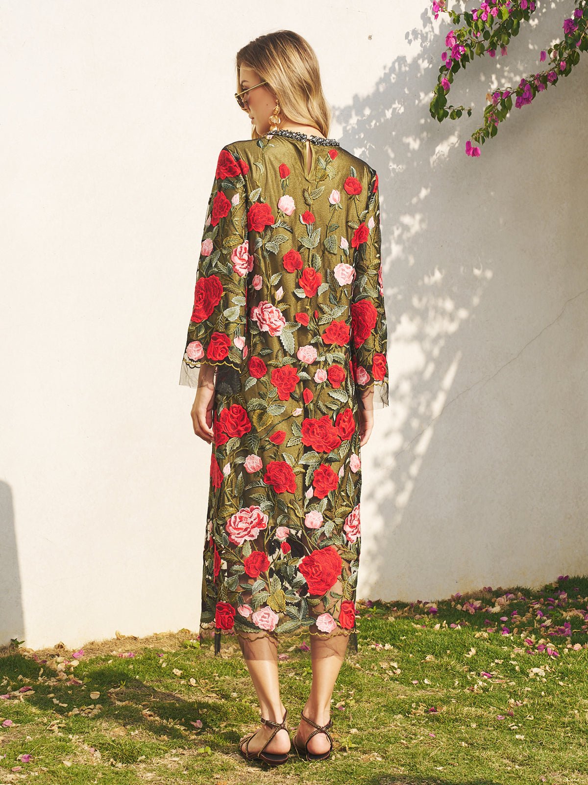 A woman stands outdoors facing a white wall, wearing the Social Queen Rose Embroidered Beaded A-Line Dress—an elegant choice for a wedding guest—and sandals.