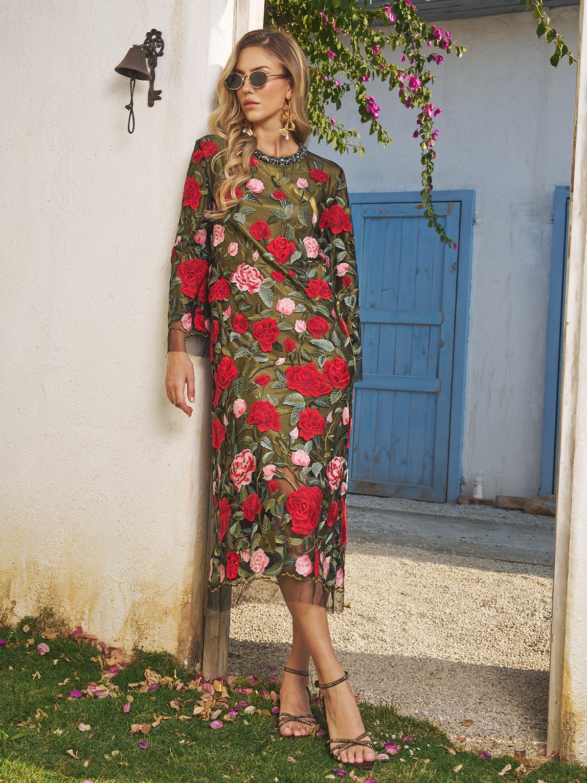 A woman wearing the Social Queen Rose Embroidered Beaded A-Line Dress and sunglasses stands outdoors by a white wall with blue doors, leaning against a column surrounded by flowers—an elegant choice for any stylish wedding guest.
