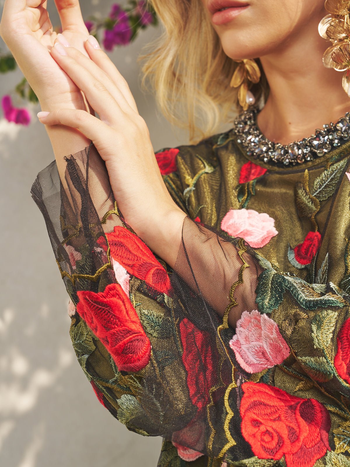 A woman wears the Social Queen Rose Embroidered Beaded A-Line Dress with sheer sleeves, large gold earrings, and pink flowers in the background—a chic choice for a stylish wedding guest.