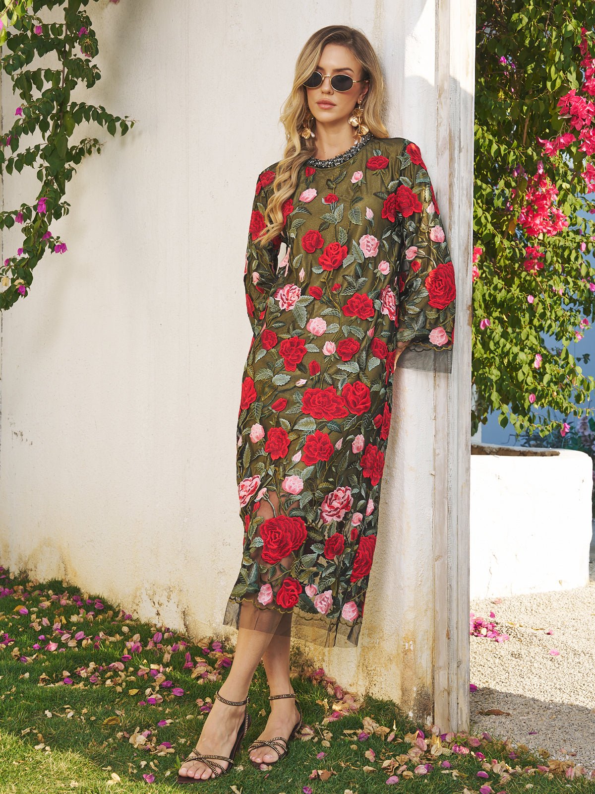 A woman in the Social Queen Rose Embroidered Beaded A-Line Dress, paired with sunglasses and sandals, stands by a white wall with floral greenery—a chic look for any stylish wedding guest.