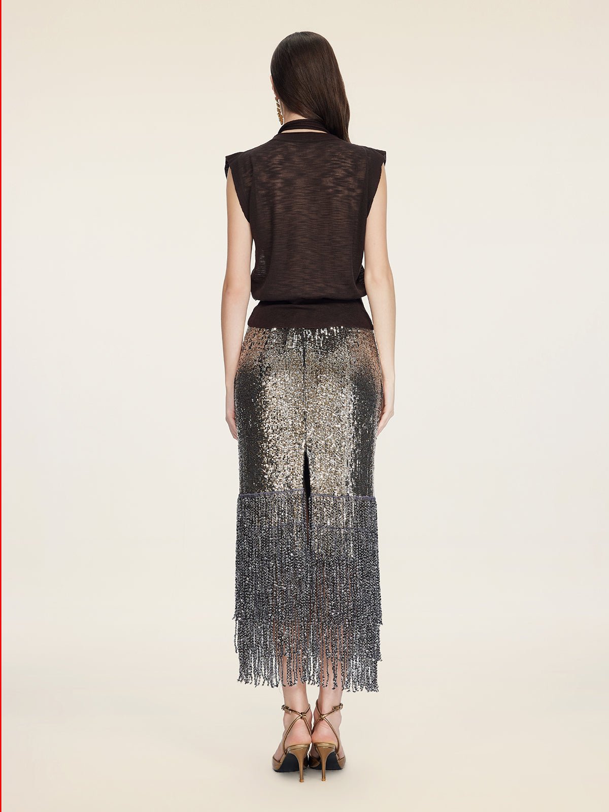 A woman stands with her back to the camera in a Social Queen Rose-Tie Neck Knit Cardigan and a silver sequin skirt with fringe, set against a plain beige background.
