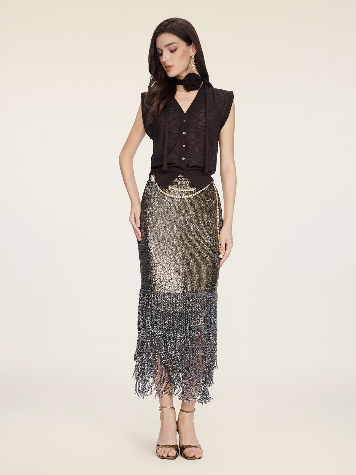 A woman wears the Social Queen Rose-Tie Neck Knit Cardigan, paired with a metallic sequined fringe skirt, gold heels, and a black scarf against a plain light background.