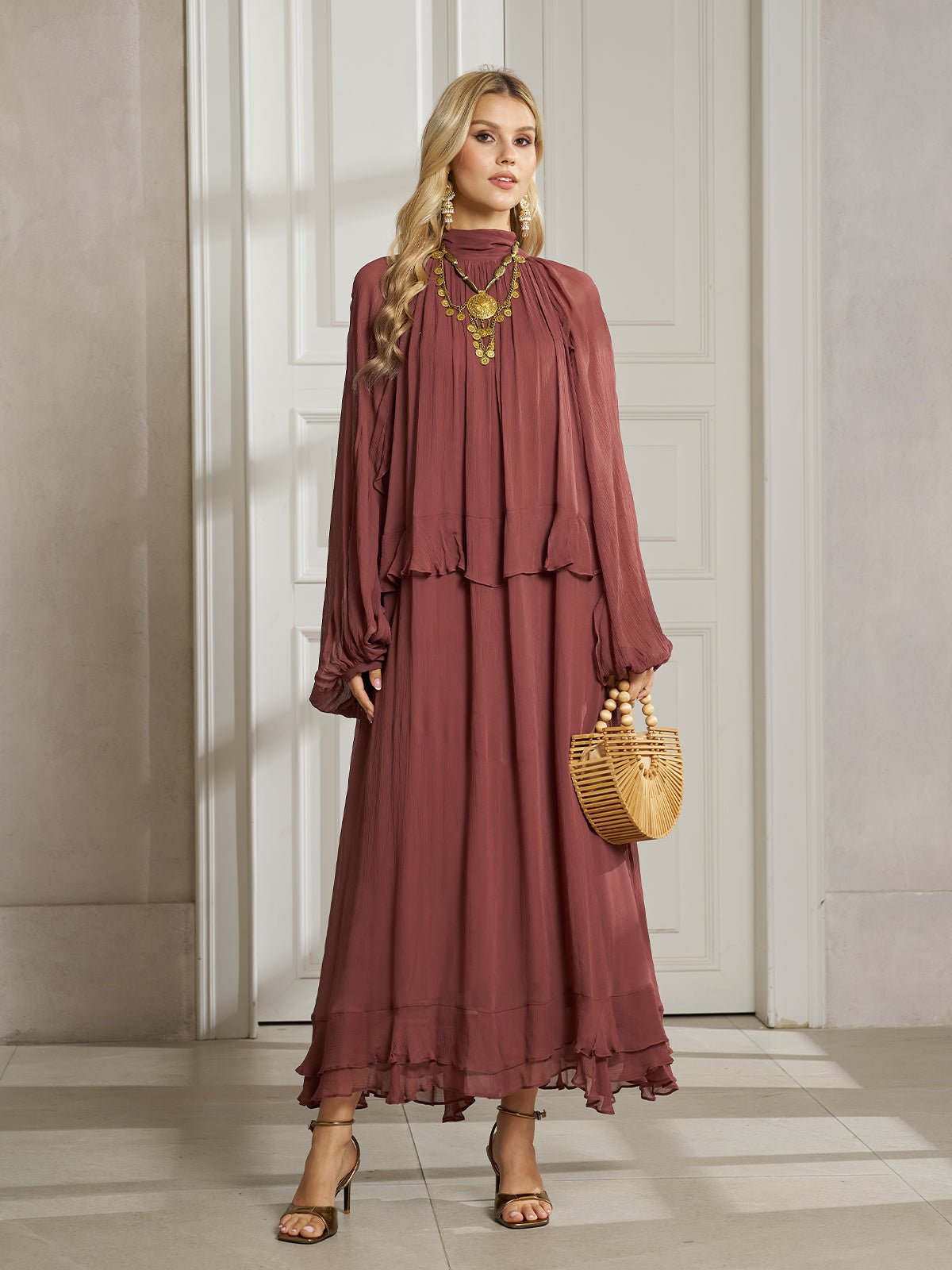 A woman stands indoors in a Social Queen Ruched High-Neck Ruffled Midi Dress with lantern sleeves and layered ruffle hem, accessorized with gold jewelry and a bamboo handbag, in front of a white-paneled door.