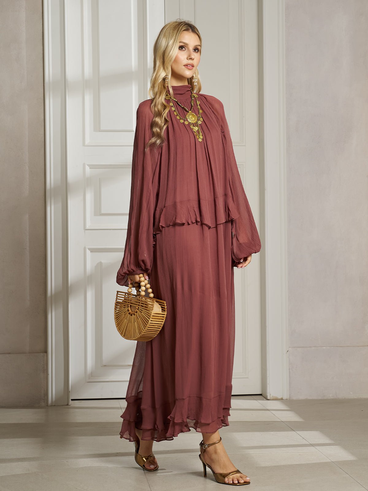 A woman stands indoors in the Social Queen Ruched High-Neck Ruffled Midi Dress, accessorized with a wooden handbag, heeled sandals, and a statement necklace.