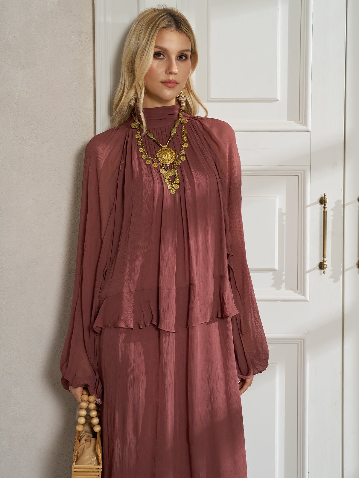 A woman wears the Social Queen Ruched High-Neck Ruffled Midi Dress with a gold statement necklace and holds a beige handbag indoors.