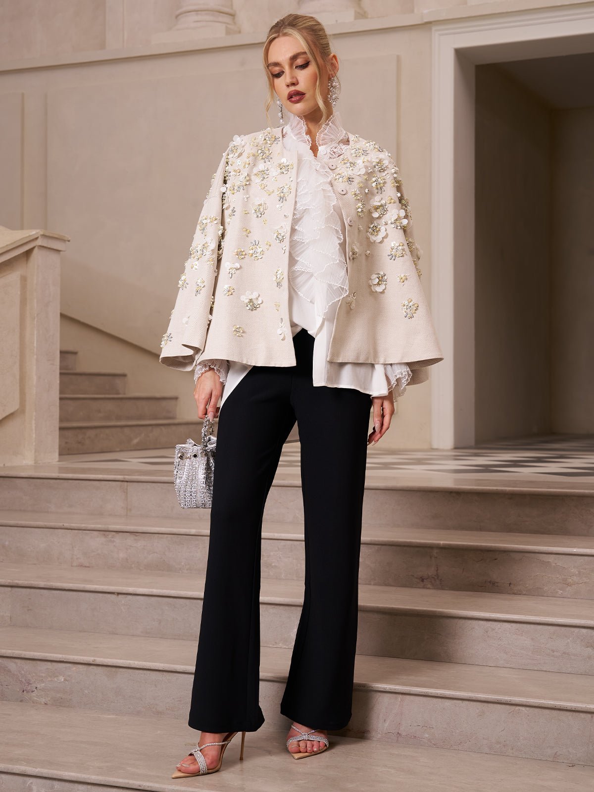 A woman stands on marble stairs wearing the Ruffled Lace Blouse with Asymmetric Hem by Social Queen under a cream embellished jacket, paired with black trousers, a small silver bag, and heeled sandals.
