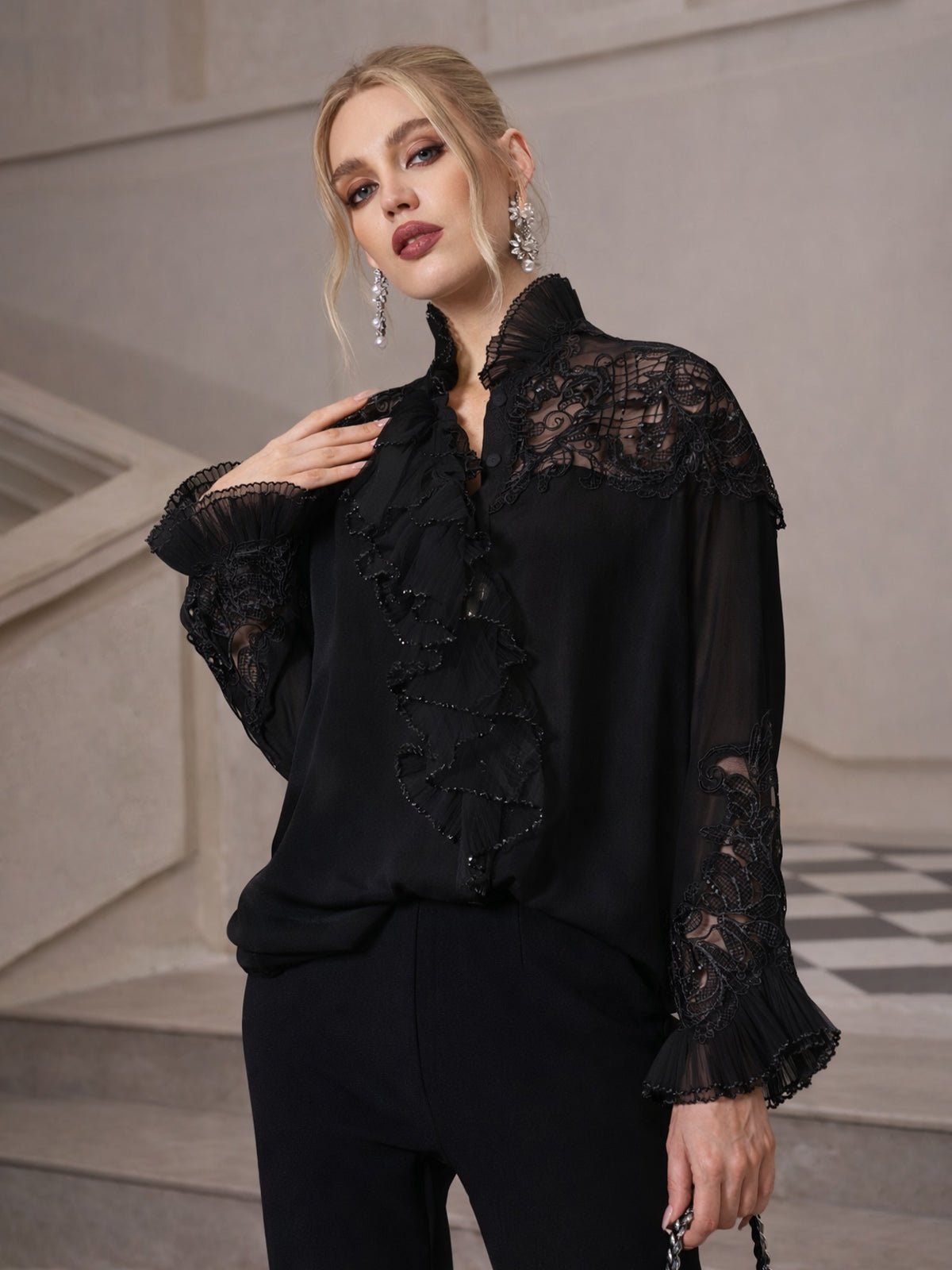 A woman stands indoors wearing the Social Queen Ruffled Lace Blouse with Asymmetric Hem and black pants, posing with one hand near her collar.