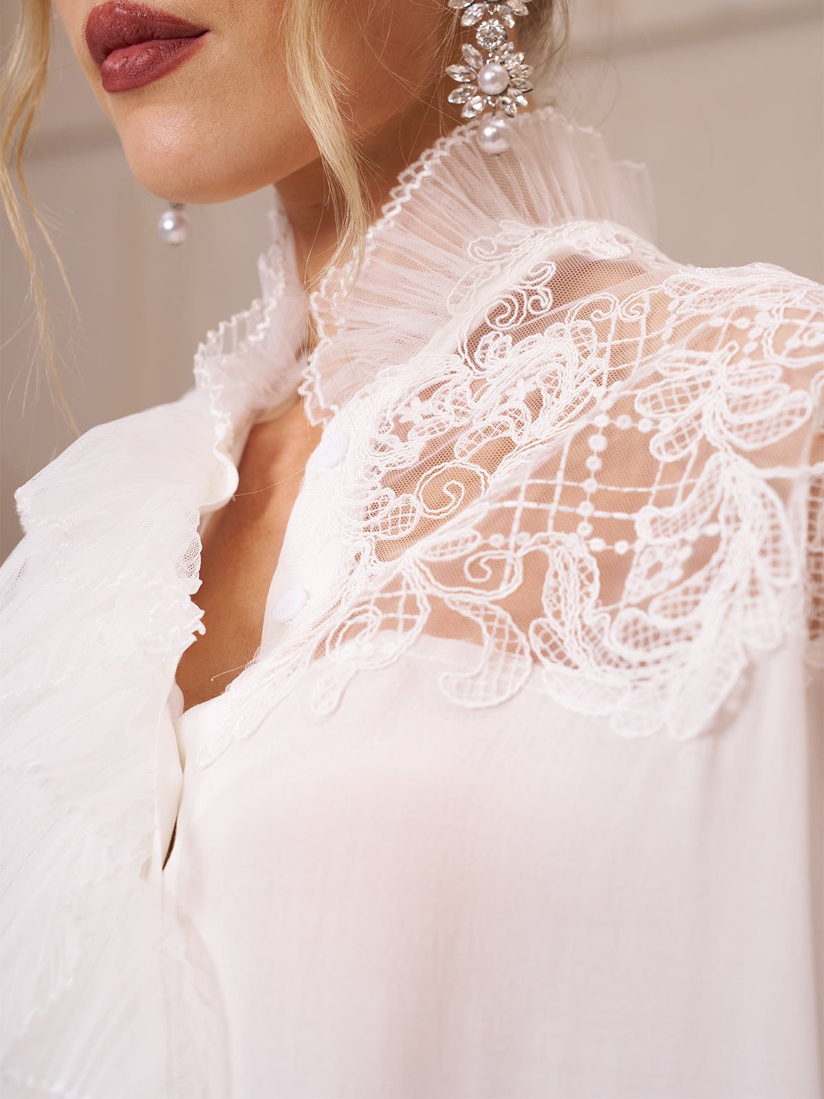 Close-up of a woman wearing the Social Queen Ruffled Lace Blouse with Asymmetric Hem, featuring organza ruffles at the collar and pearl earrings; only part of her face and upper body are visible.