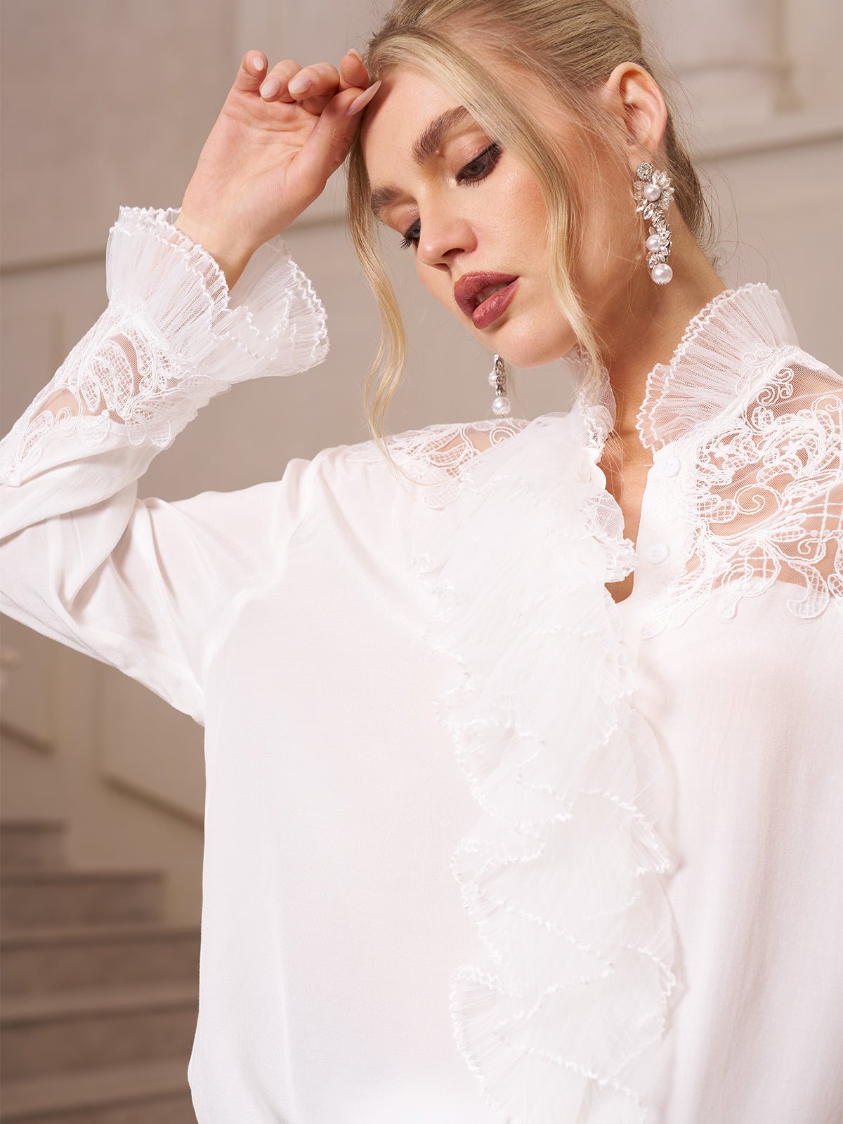 A woman indoors wears the Social Queen Ruffled Lace Blouse with Asymmetric Hem, featuring delicate organza ruffles; she touches her forehead and looks downward.