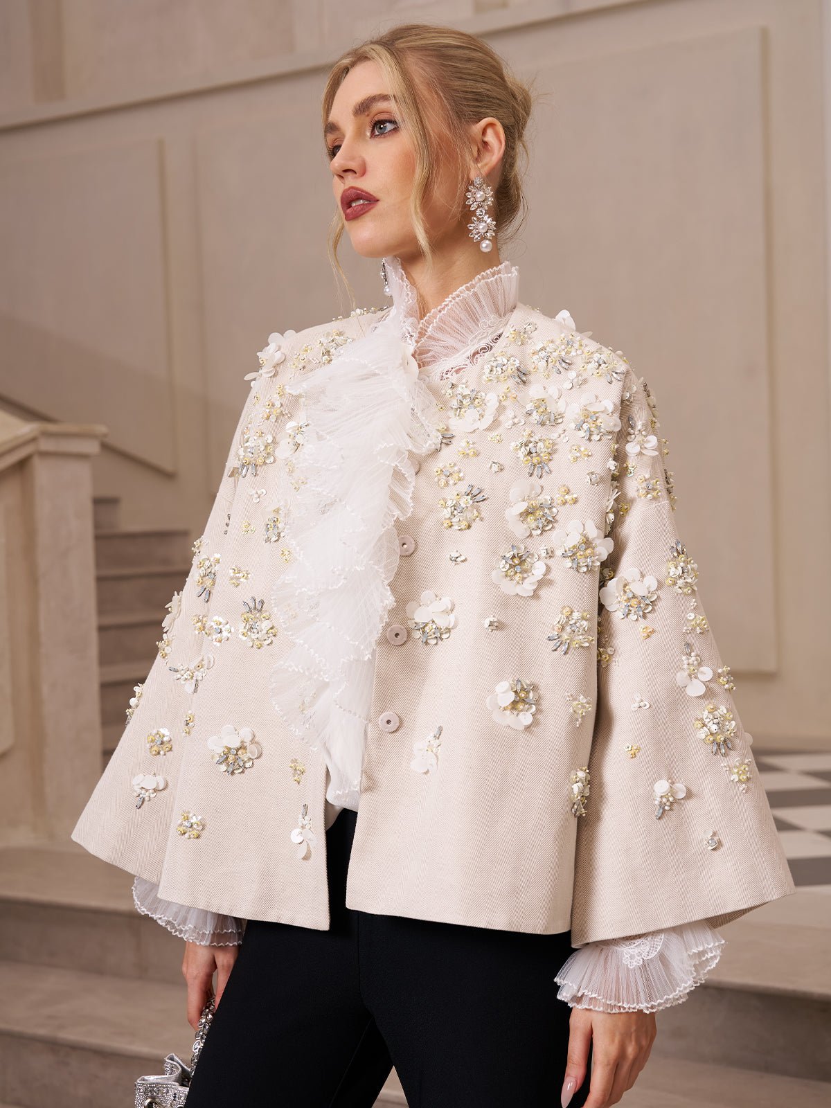 Woman indoors wears Social Queen’s Ruffled Lace Blouse with Asymmetric Hem, featuring cream fabric, floral embellishments, and organza ruffles. She pairs it with black pants, drop earrings, and a silver clutch.