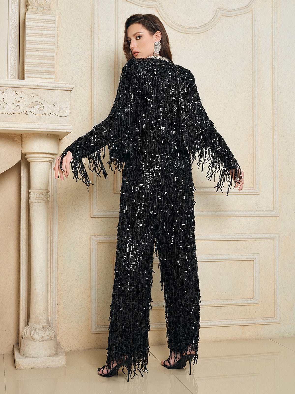 A woman poses indoors near a cream ornate wall and fireplace, wearing the Social Queen Sequin & Crystal Fringe Cardigan with matching pants and high-heeled sandals, showcasing chic eveningwear.