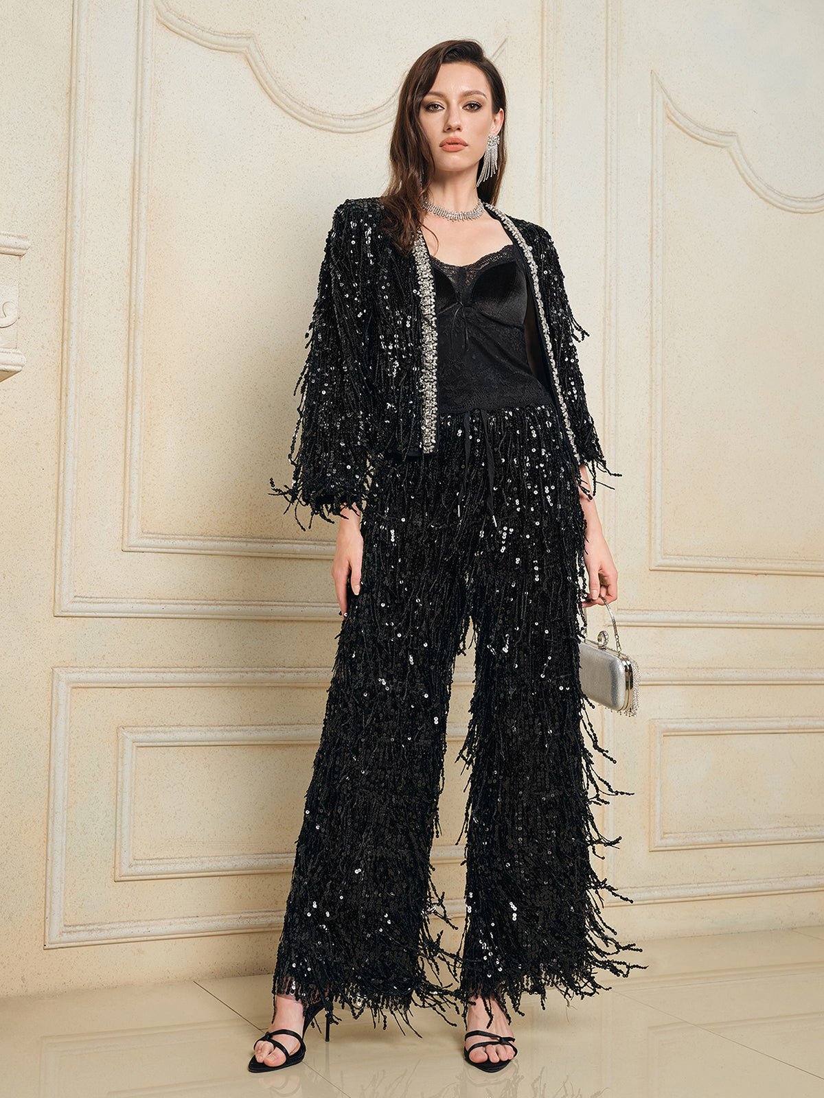 A woman stands indoors wearing the Social Queen Sequin & Crystal Fringe Cardigan with matching pants, a black top, black heeled sandals, a silver necklace, and holding a small metallic handbag.